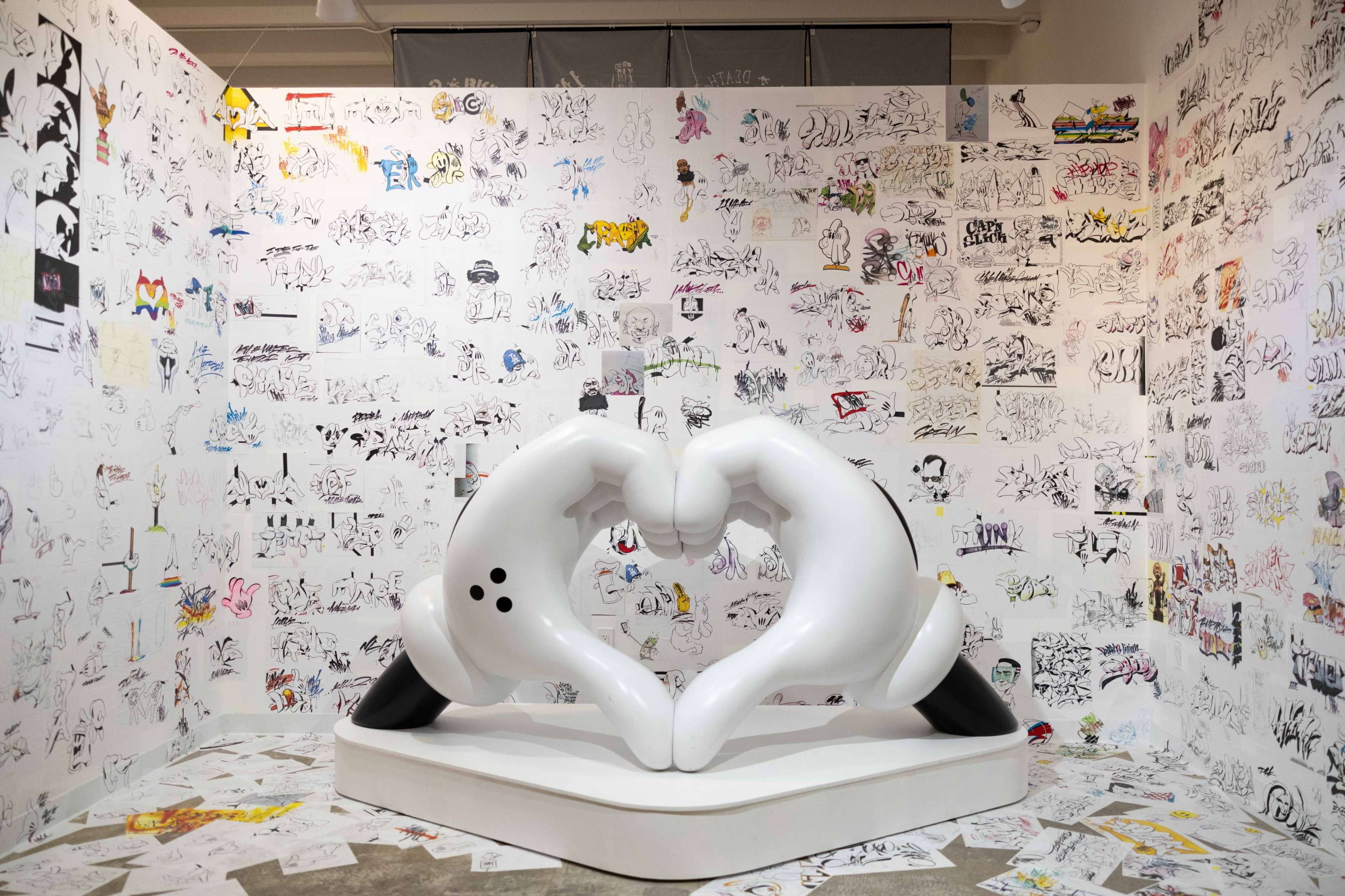 The image shows a large white sculpture of two hands forming a heart shape, surrounded by a wall covered in various black and white sketches and drawings.