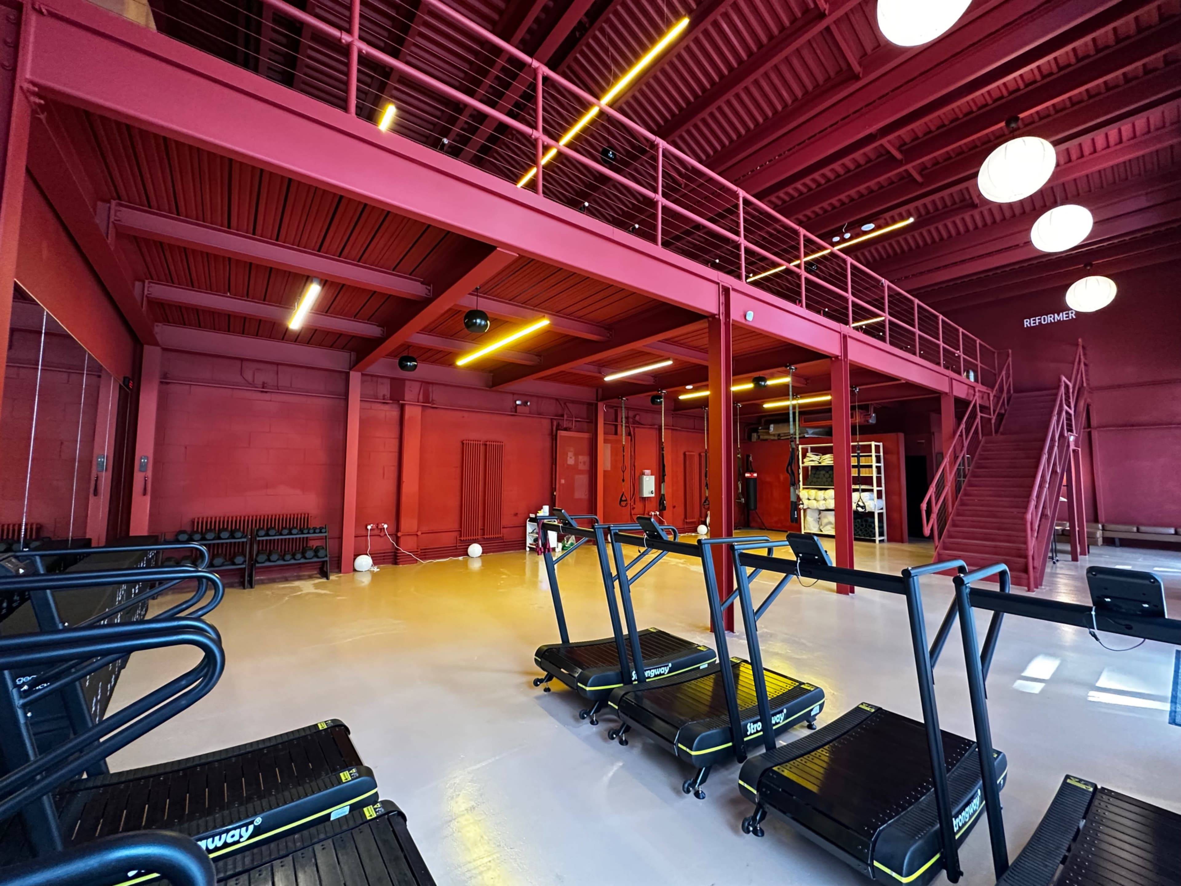 The image shows a modern fitness space with red walls, multiple exercise machines, and a staircase leading to an upper level.