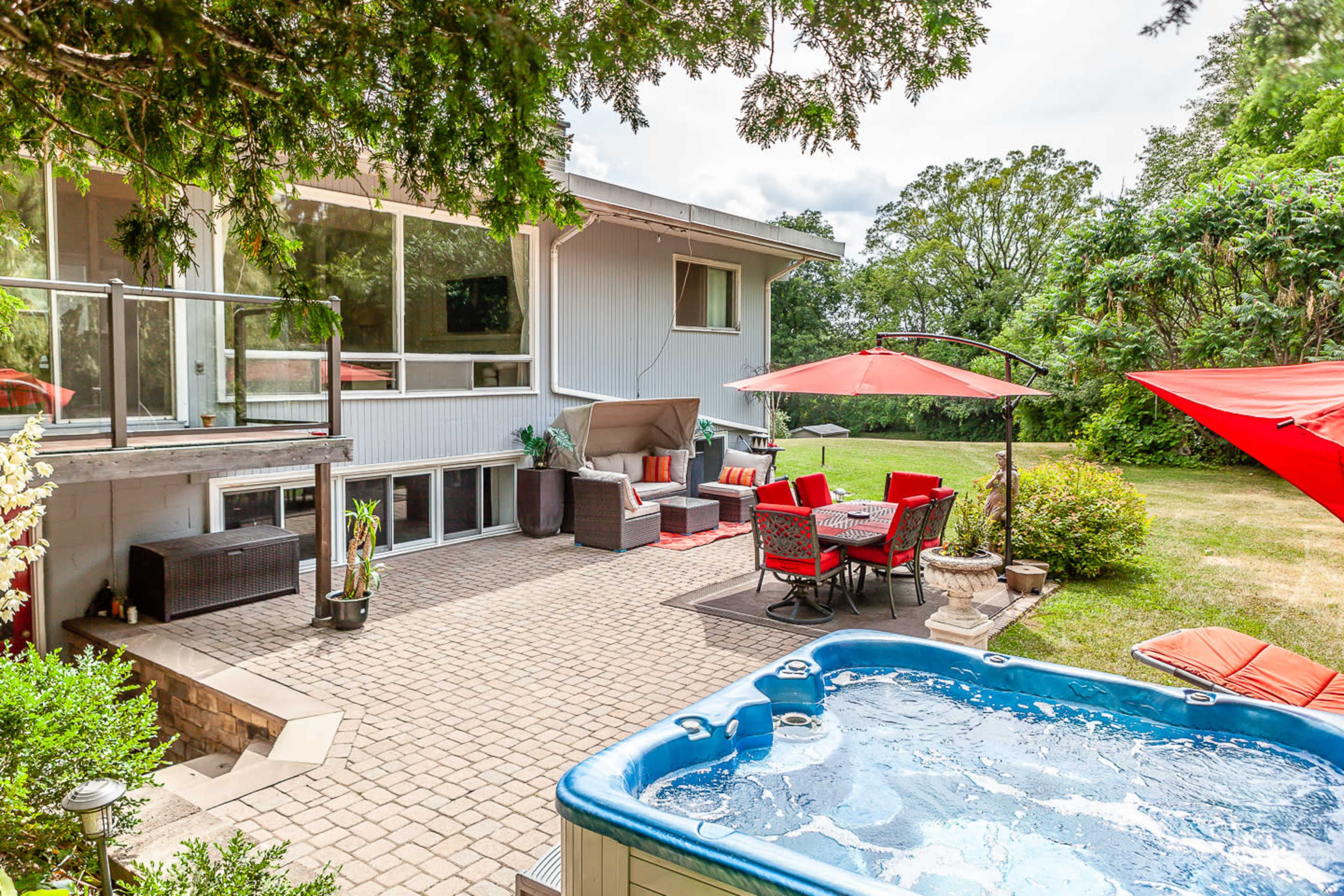 A backyard patio with a hot tub, seating area, and a dining set under red umbrellas, surrounded by greenery.