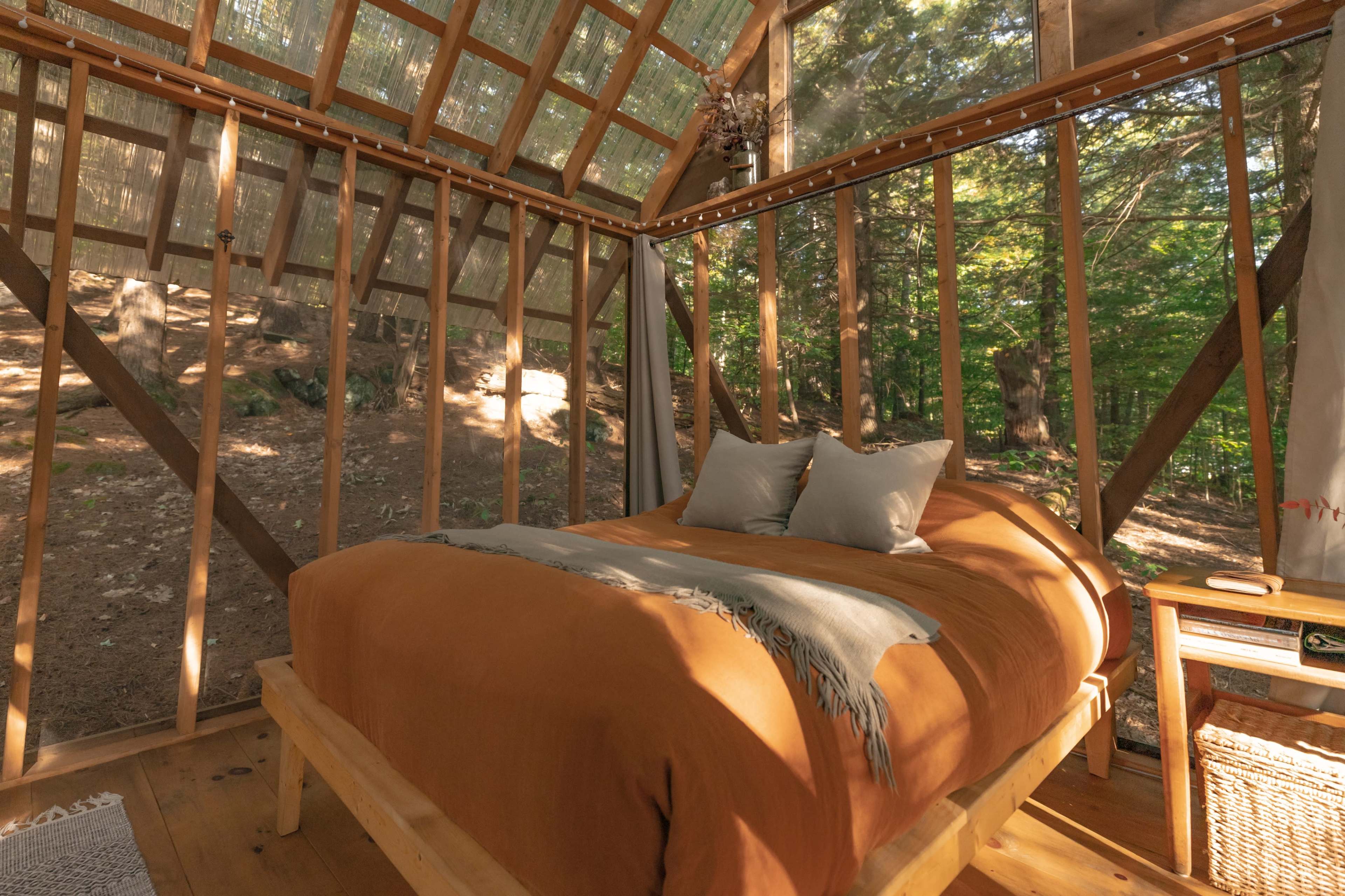 A wooden interior of a cabin with a bed draped in an orange blanket, surrounded by large windows that offer a view of a forested area.
