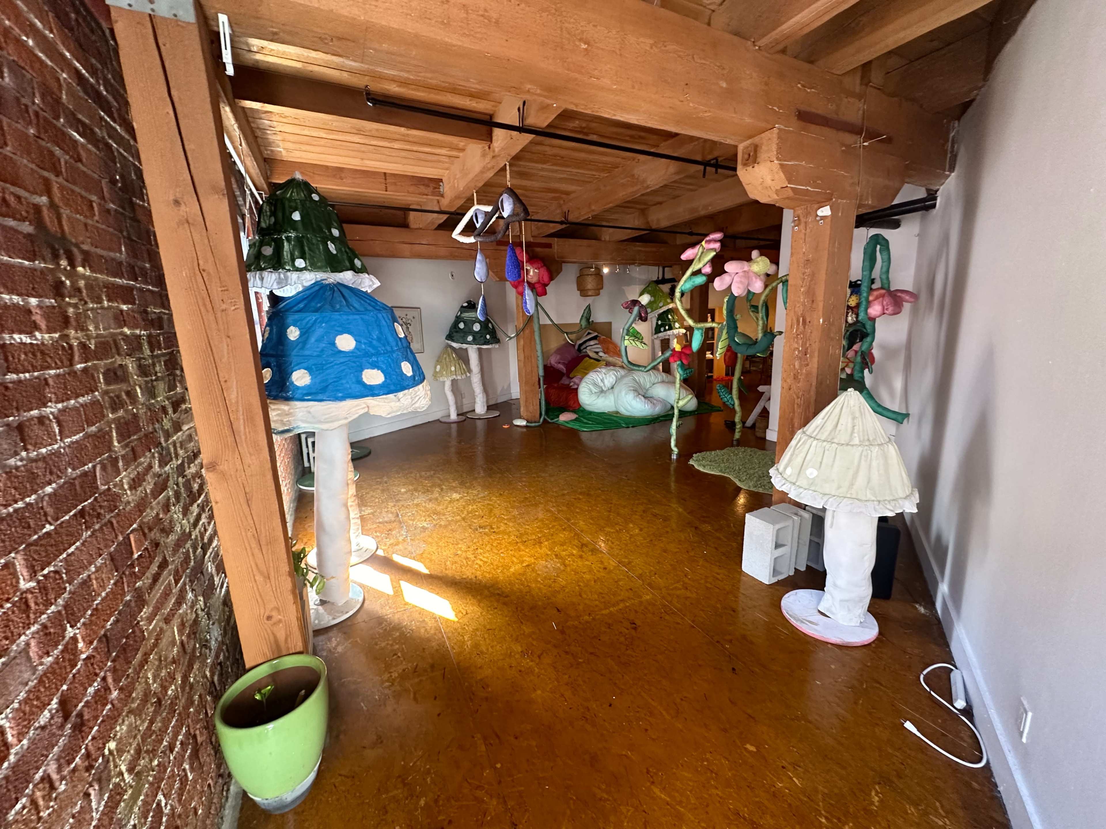 The image shows a spacious room with wooden beams, featuring colorful, oversized mushroom-shaped sculptures and whimsical decor scattered throughout.