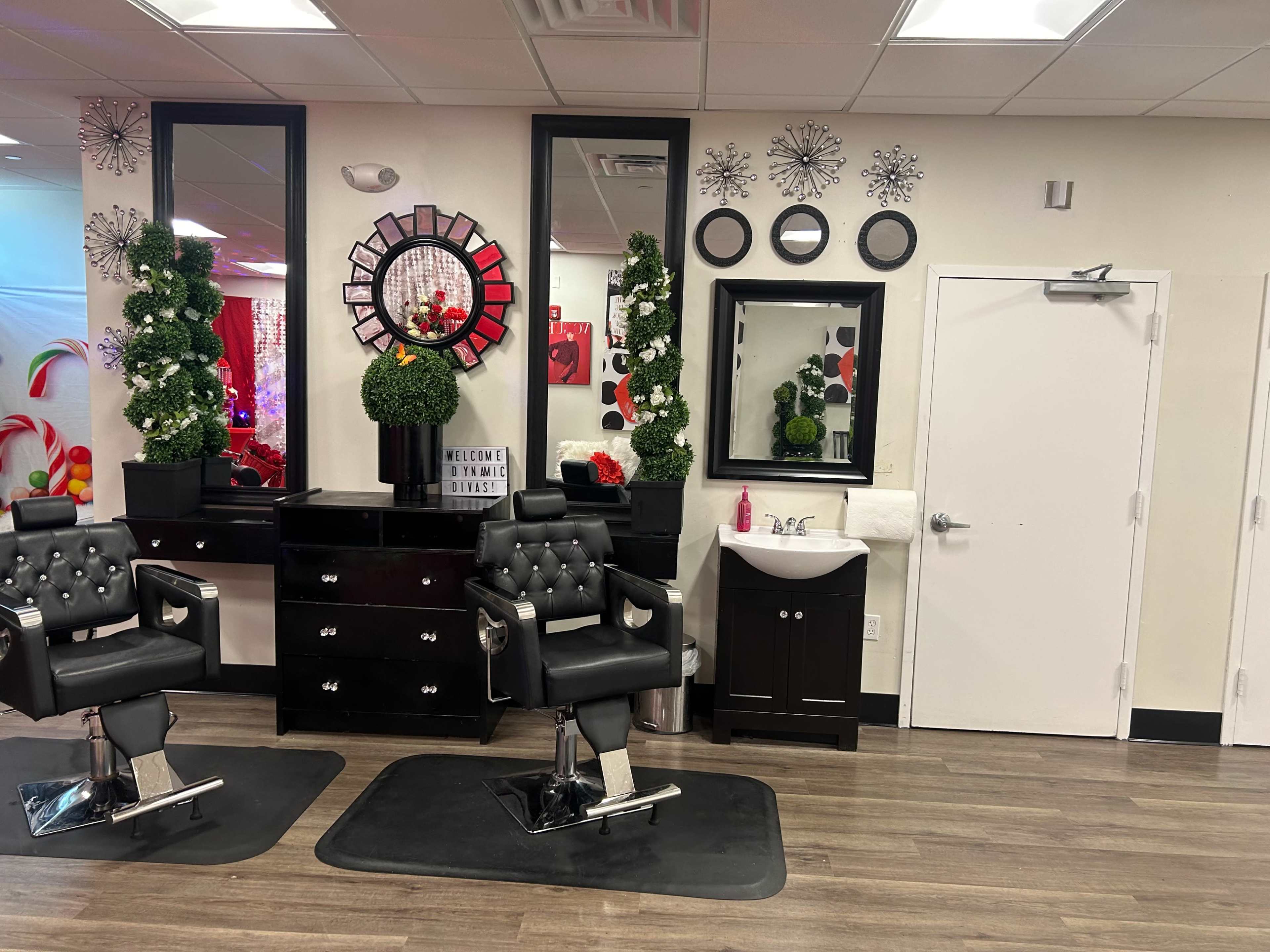 A modern salon interior with two black salon chairs, a mirrored wall, decorative plants, and a wall-mounted sink.
