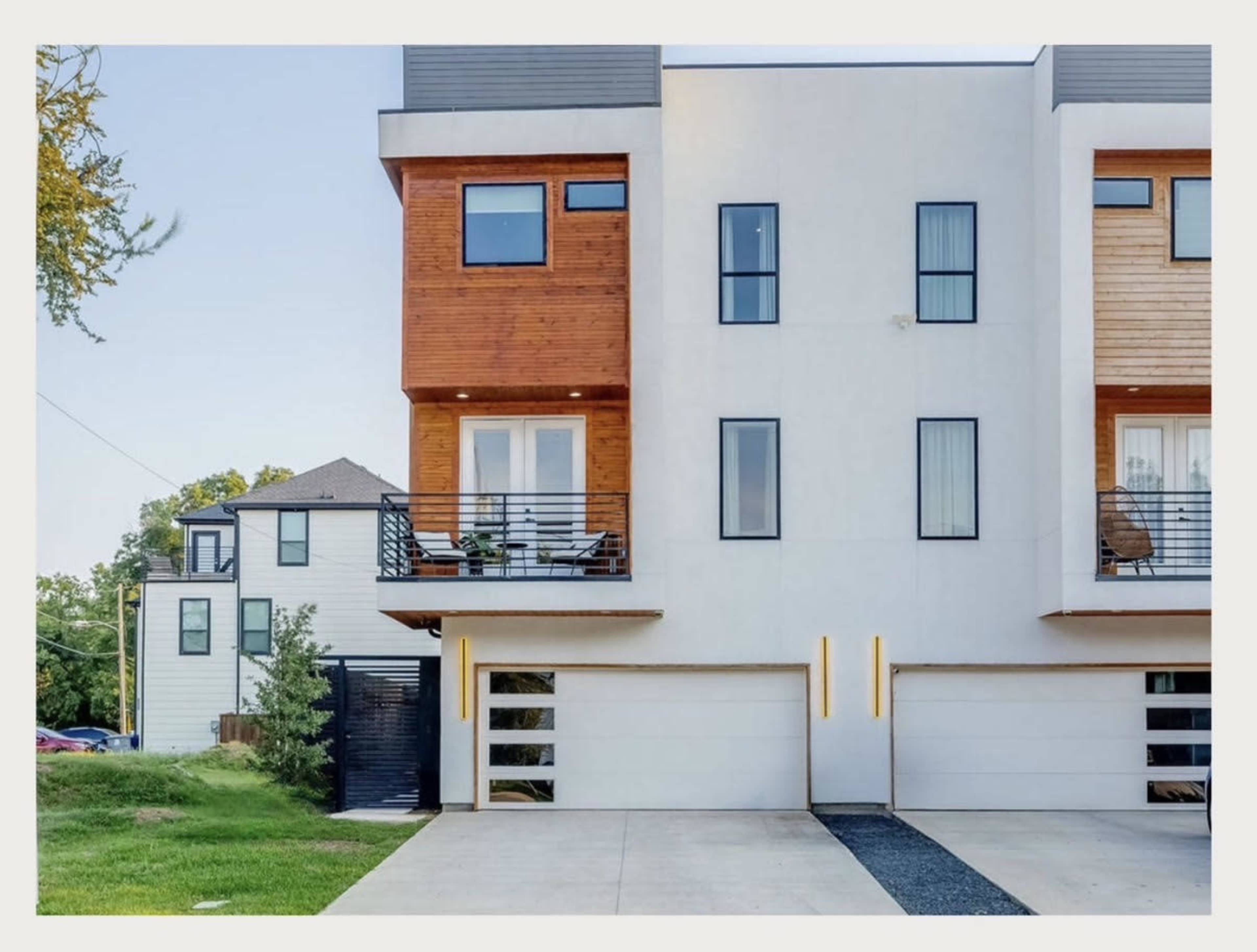 The image shows a modern, two-story townhouse with a combination of white and wooden exterior features, including a garage and balconies.