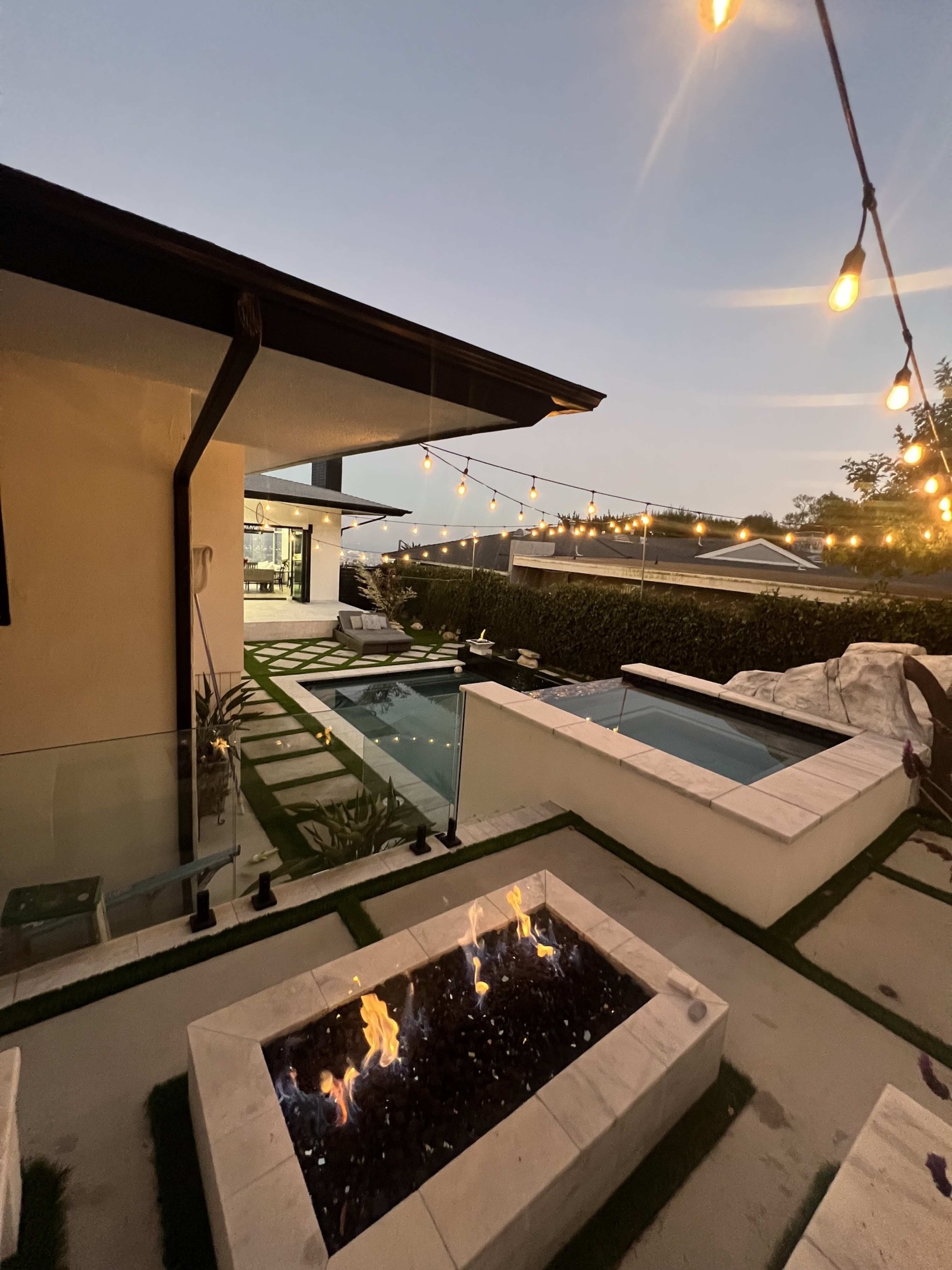 The image shows a modern backyard scene featuring a fire pit, swimming pool, and patio area lit by string lights at dusk.