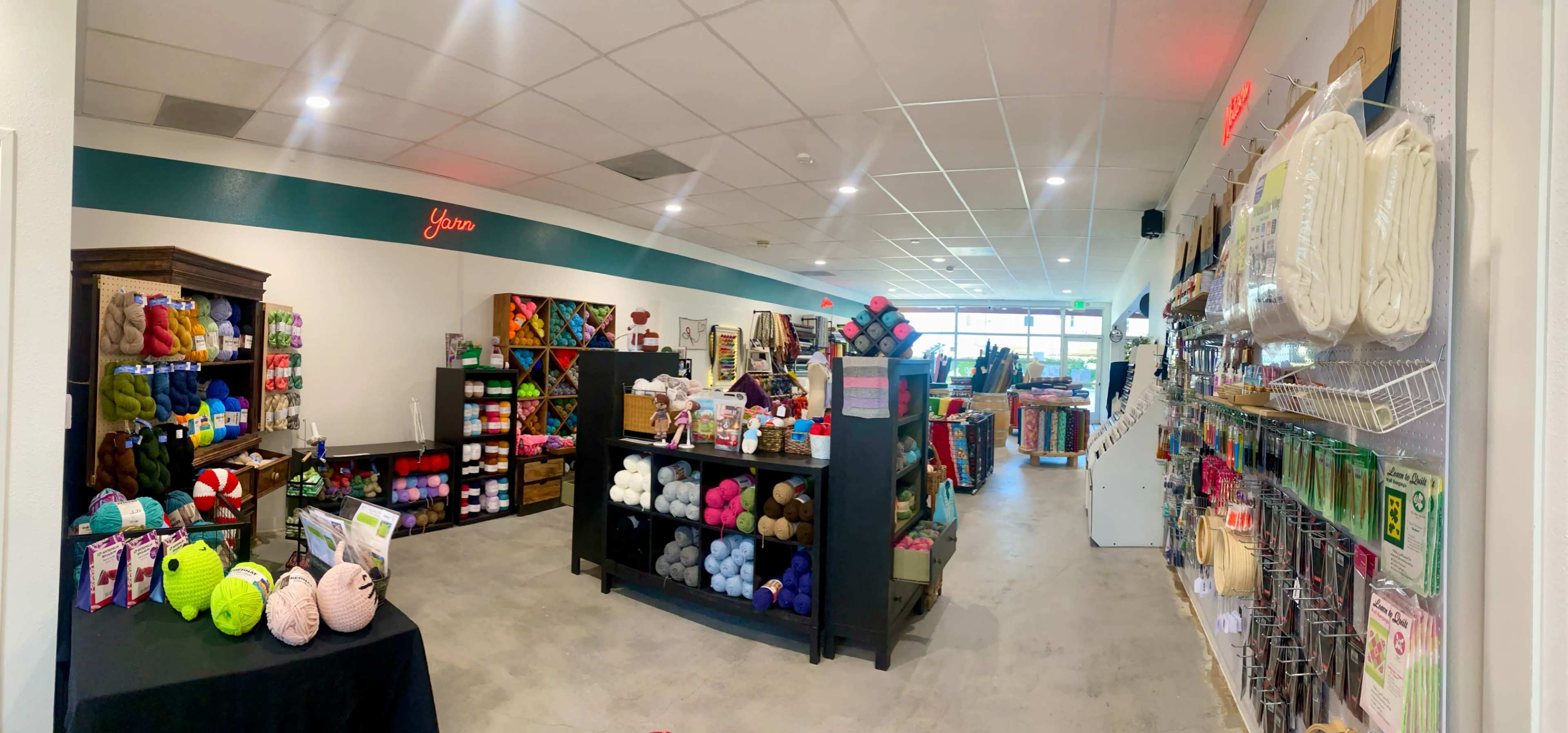 The image shows a colorful yarn and craft supply shop with shelves stocked with various types of yarn, crafting tools, and materials, all arranged in an open space.