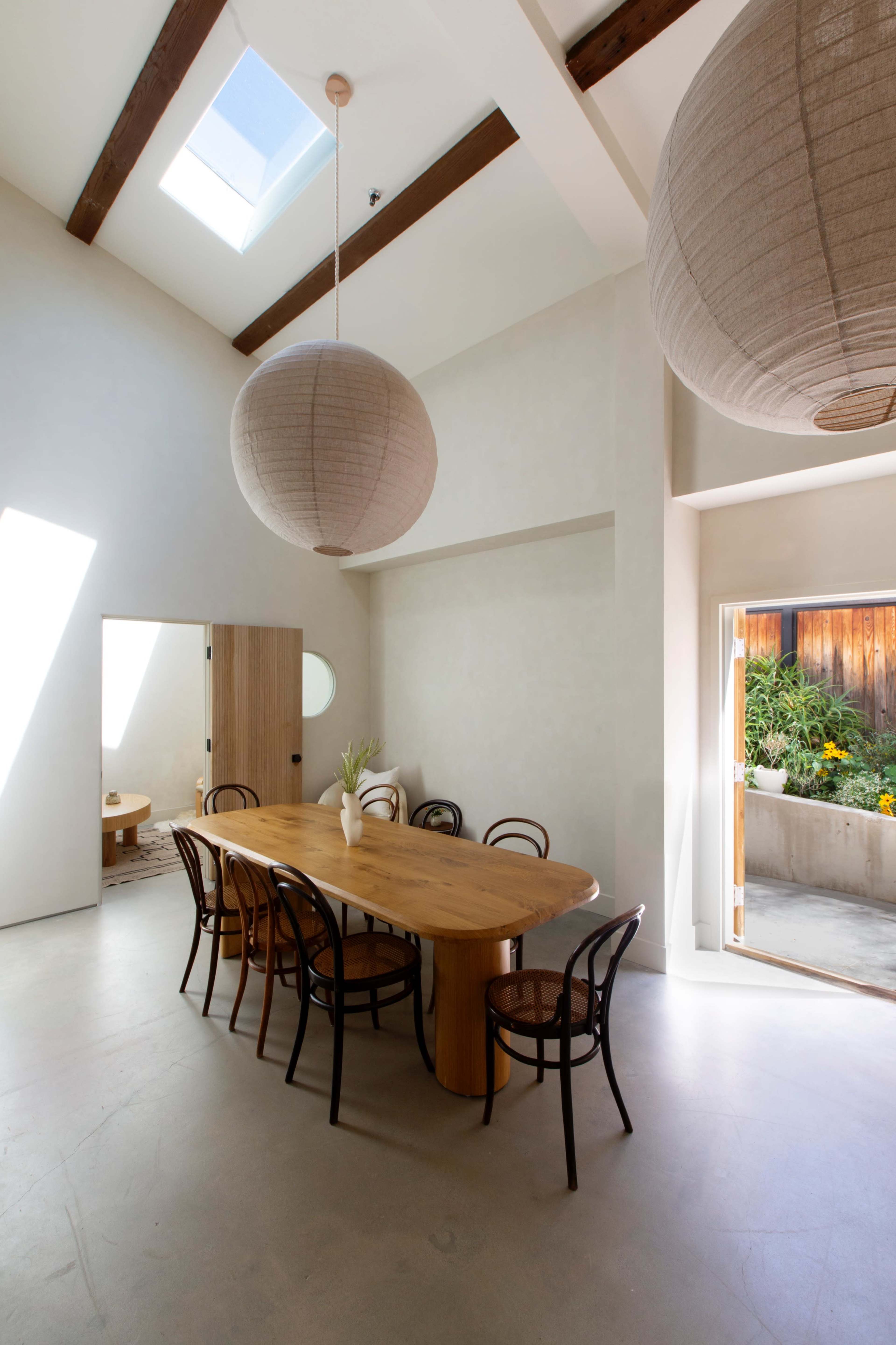 A spacious dining area features a large wooden table surrounded by curved chairs, illuminated by natural light from a skylight and large sliding door.