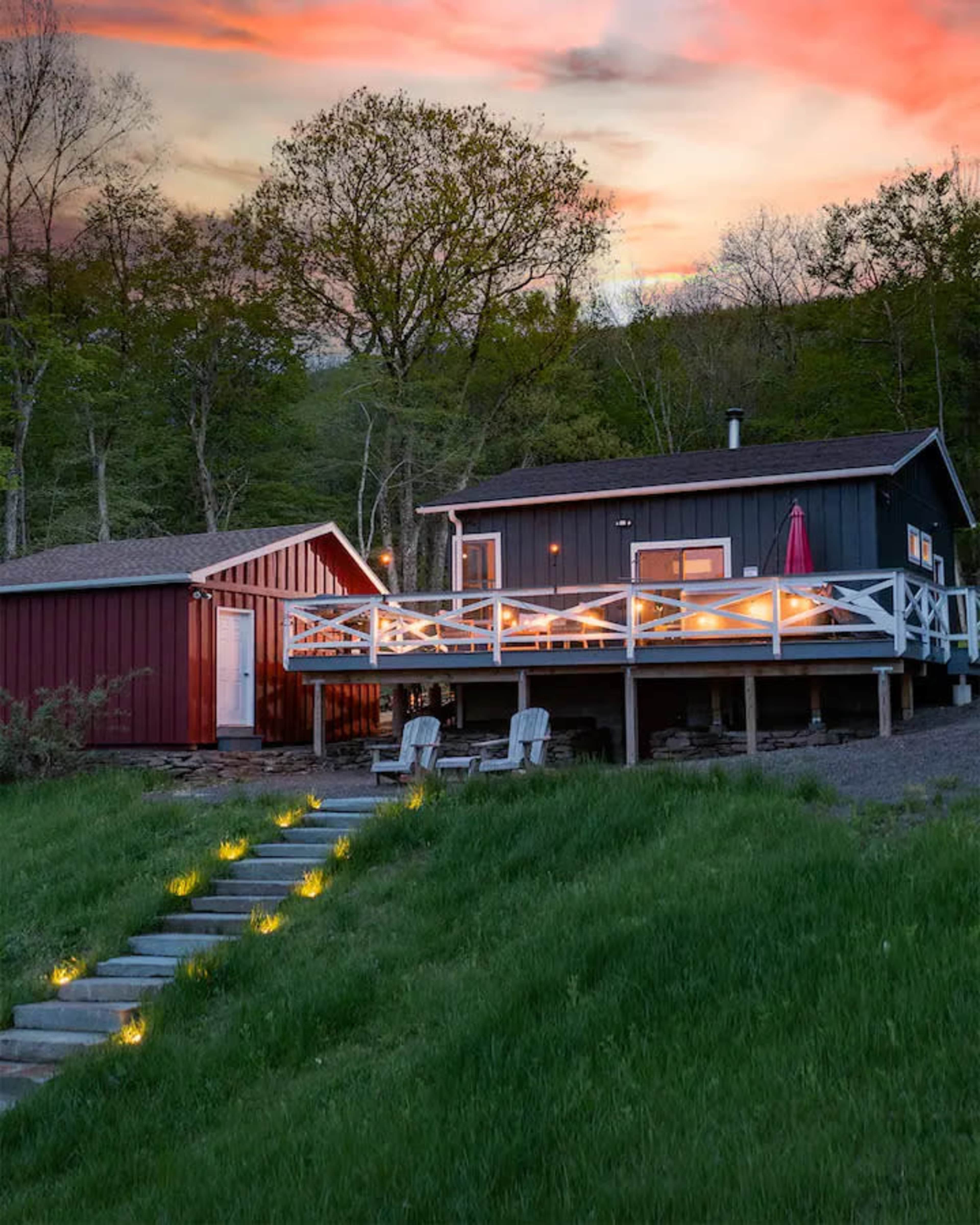 A cabin with a wooden deck and a red shed is situated on a grassy hillside at dusk, illuminated by walkway lights.