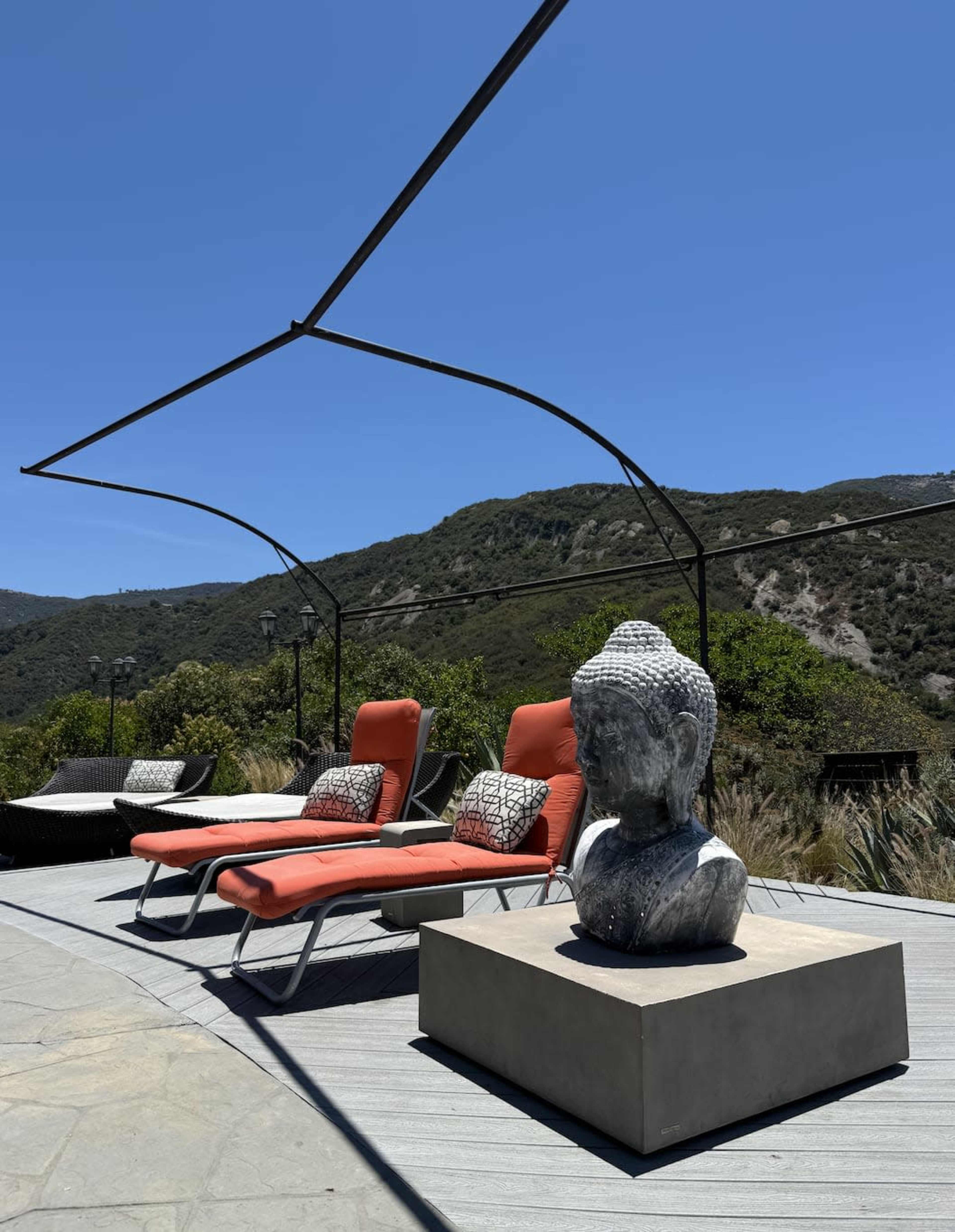 The image shows two red lounge chairs beside a concrete pedestal with a Buddha statue, set against a backdrop of green hills and a clear blue sky.