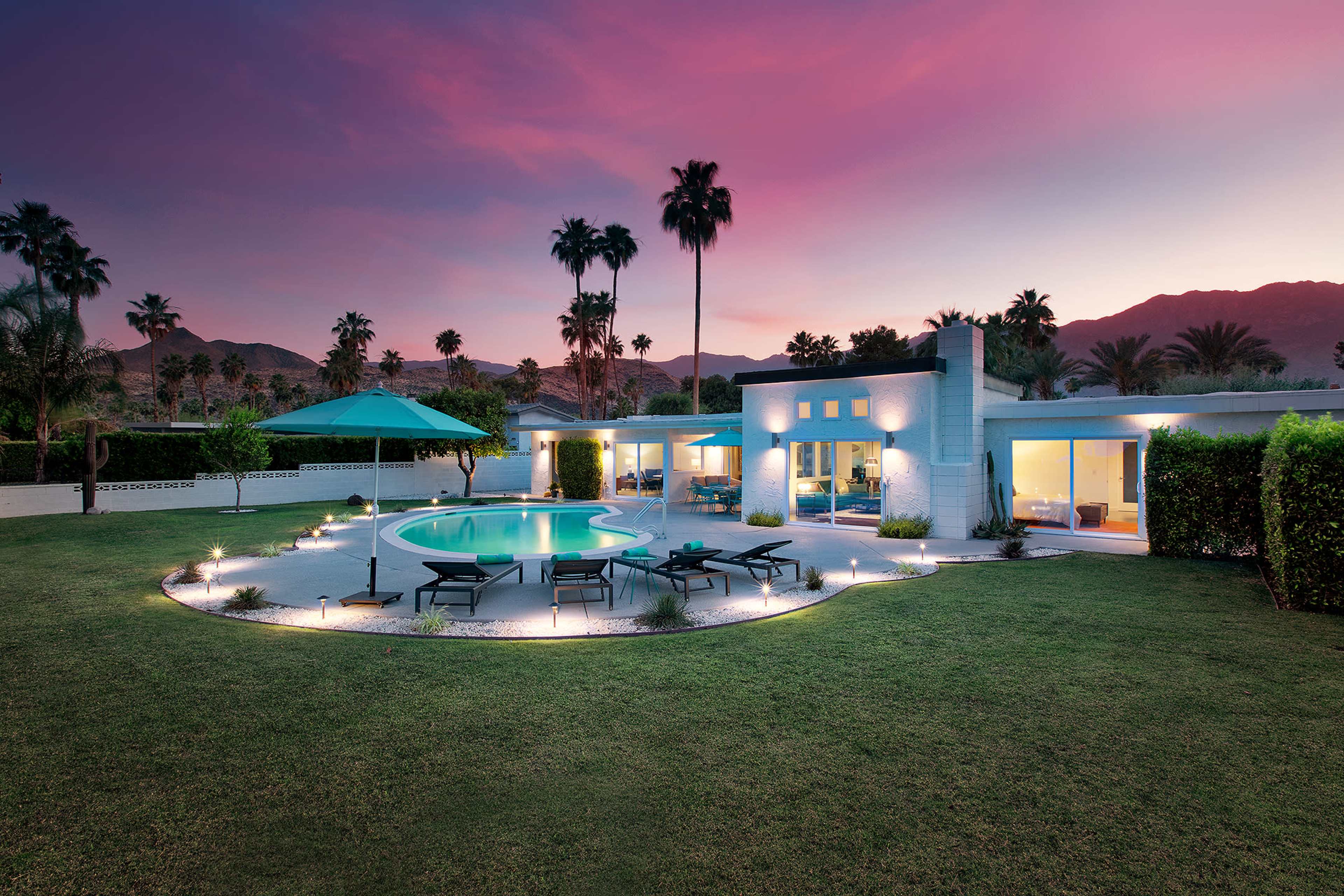 A modern house with a swimming pool and palm trees is set against a colorful sunset in a landscaped yard.
