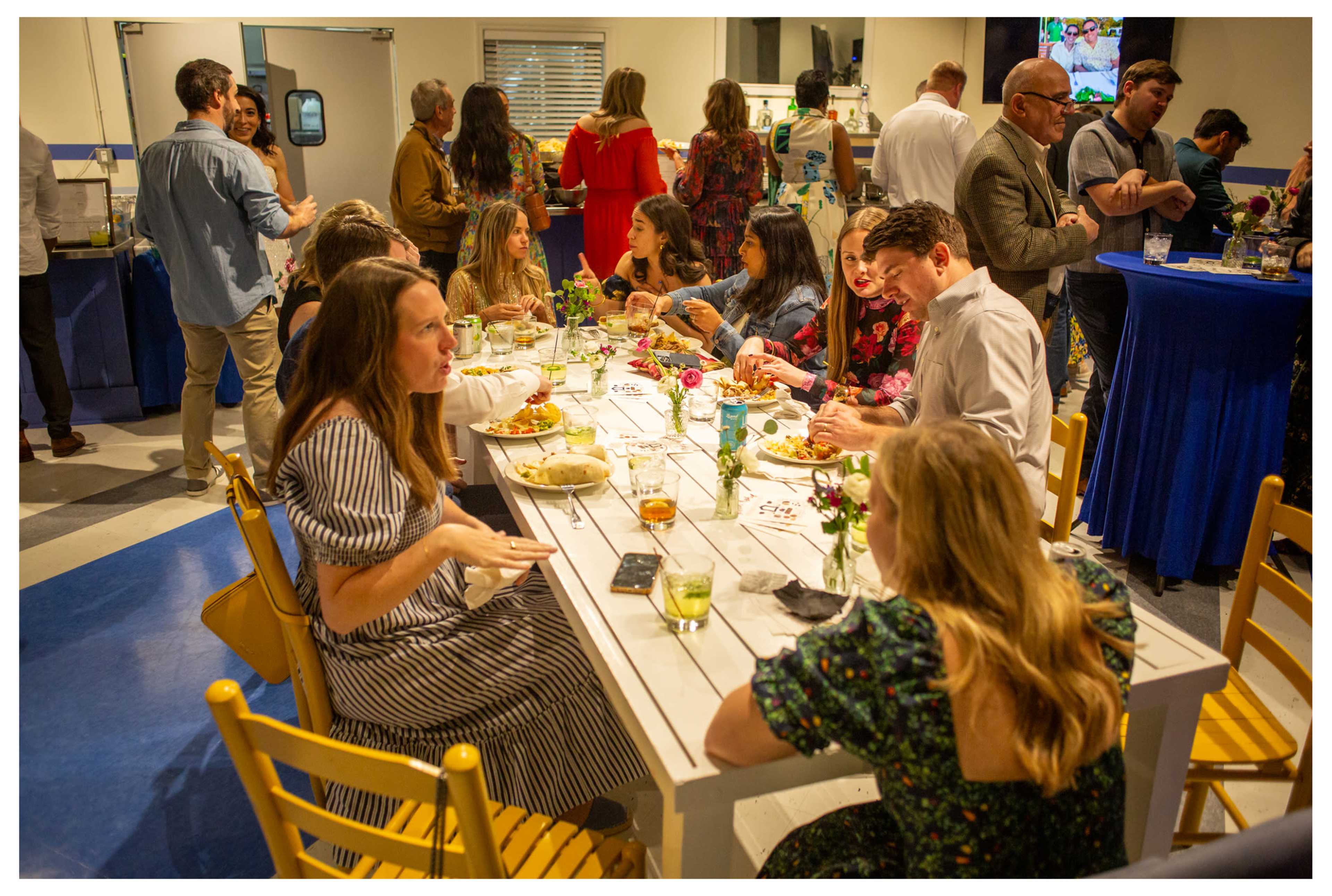 A group of people is gathered around a long table, sharing food and drinks at a lively event.