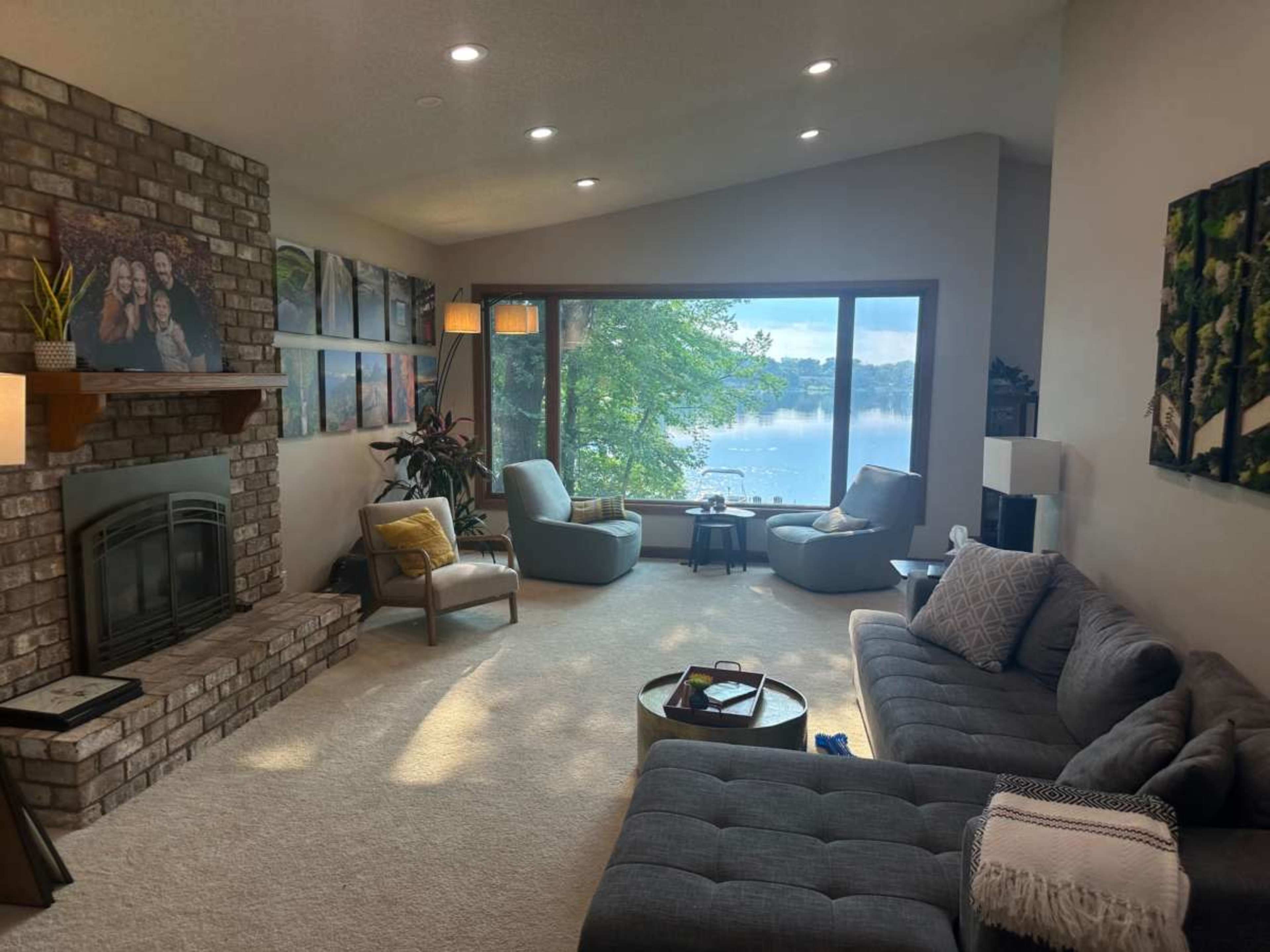 The image shows a cozy living room featuring a brick fireplace, large windows with a view of a body of water, and several comfortable seating arrangements.