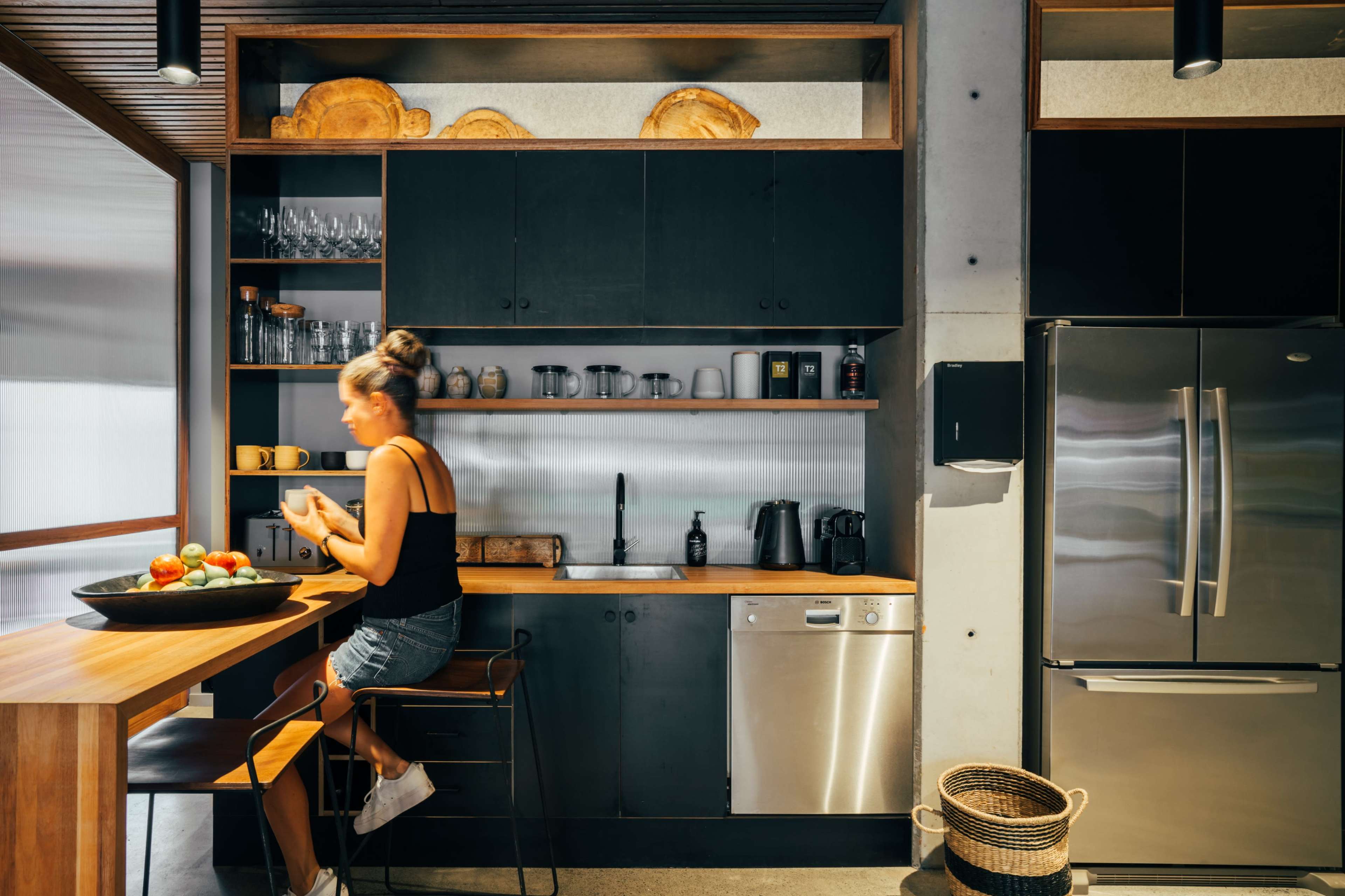 A woman sits on a high stool at a wooden counter in a modern kitchen, surrounded by neatly organized kitchenware and appliances.