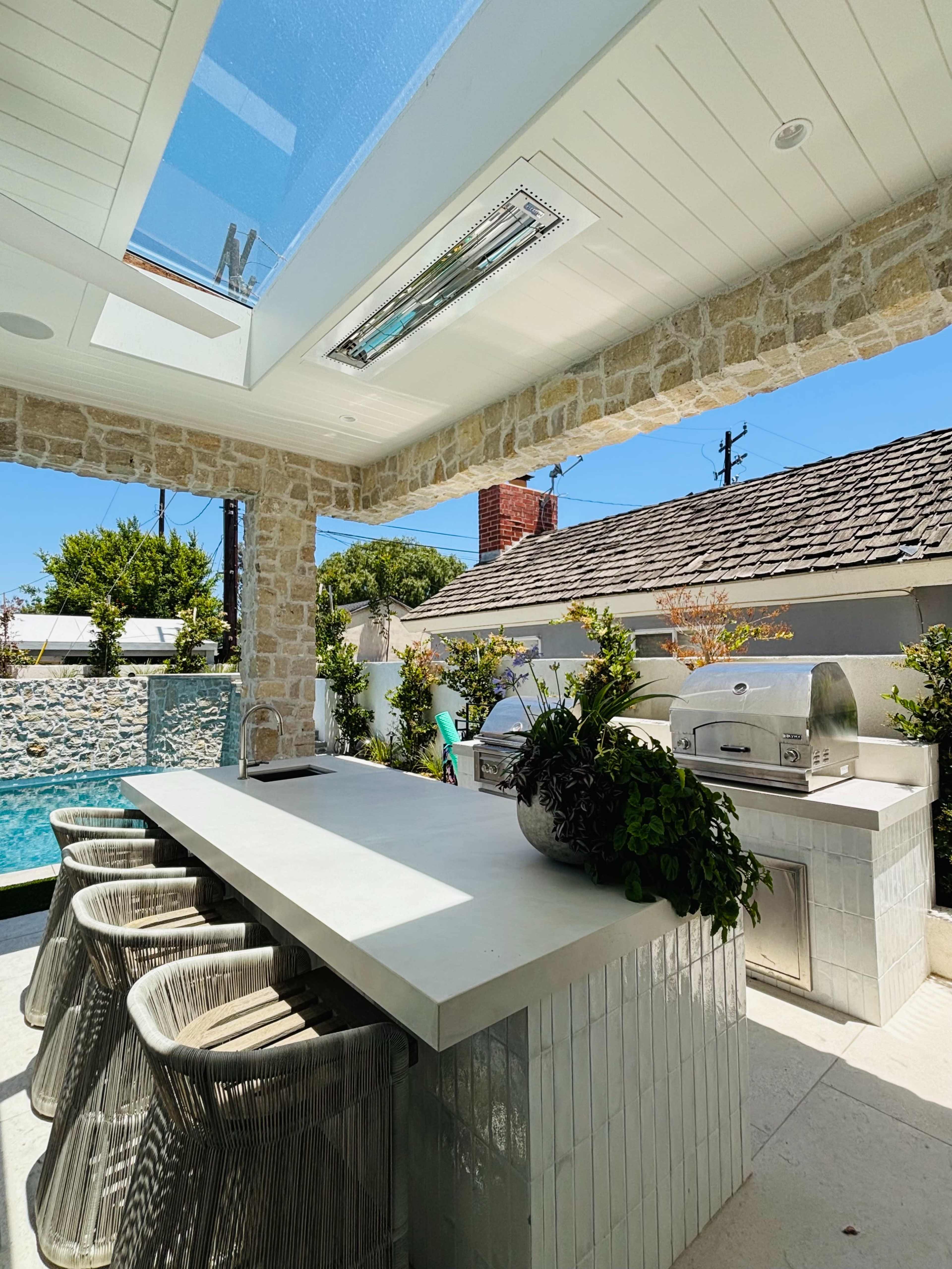 The image shows an outdoor kitchen with a large countertop, metal grills, and bar seating under a glass roof.
