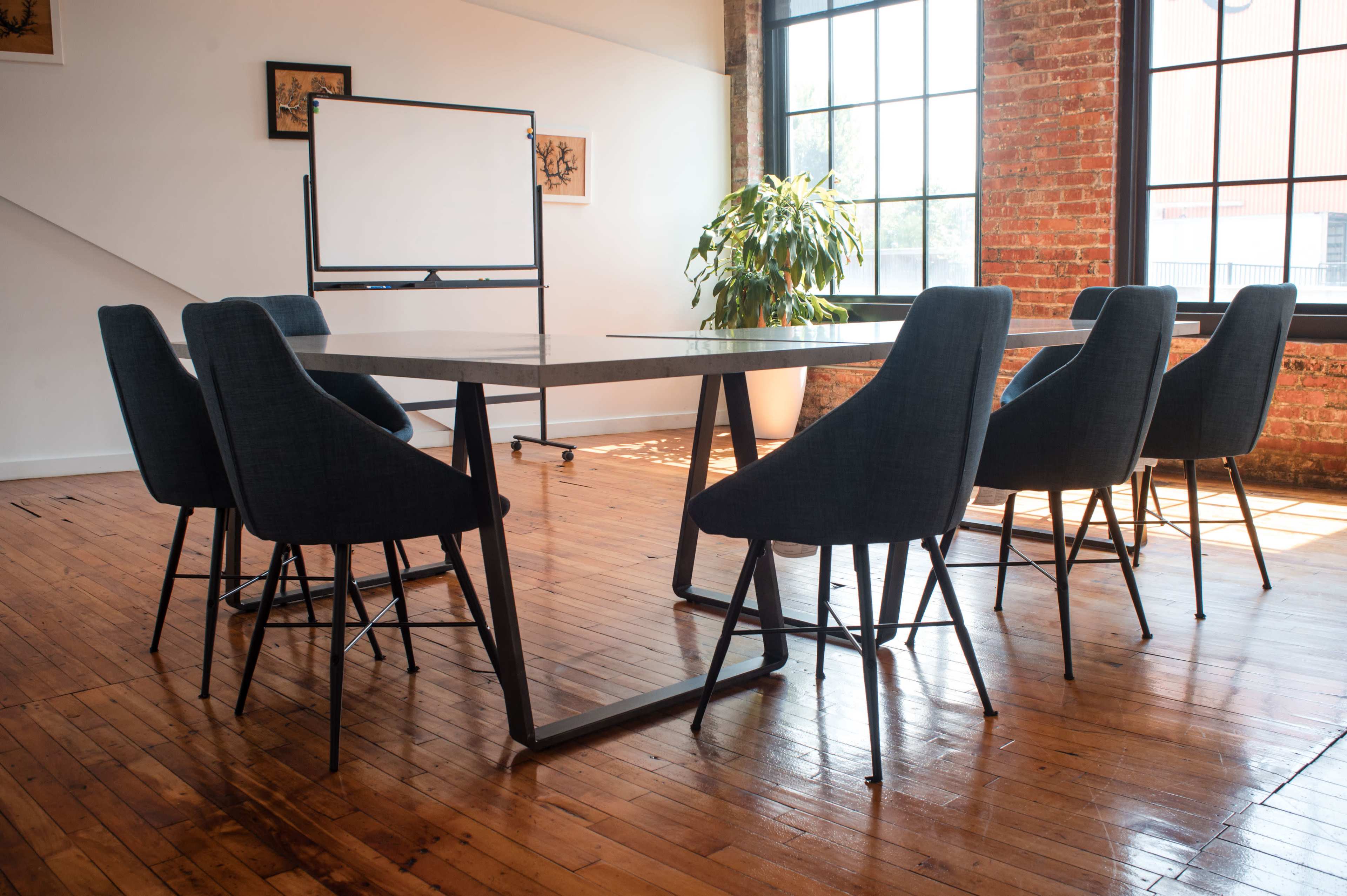 A conference room features a long table surrounded by six chairs, with large windows and a potted plant in the corner.