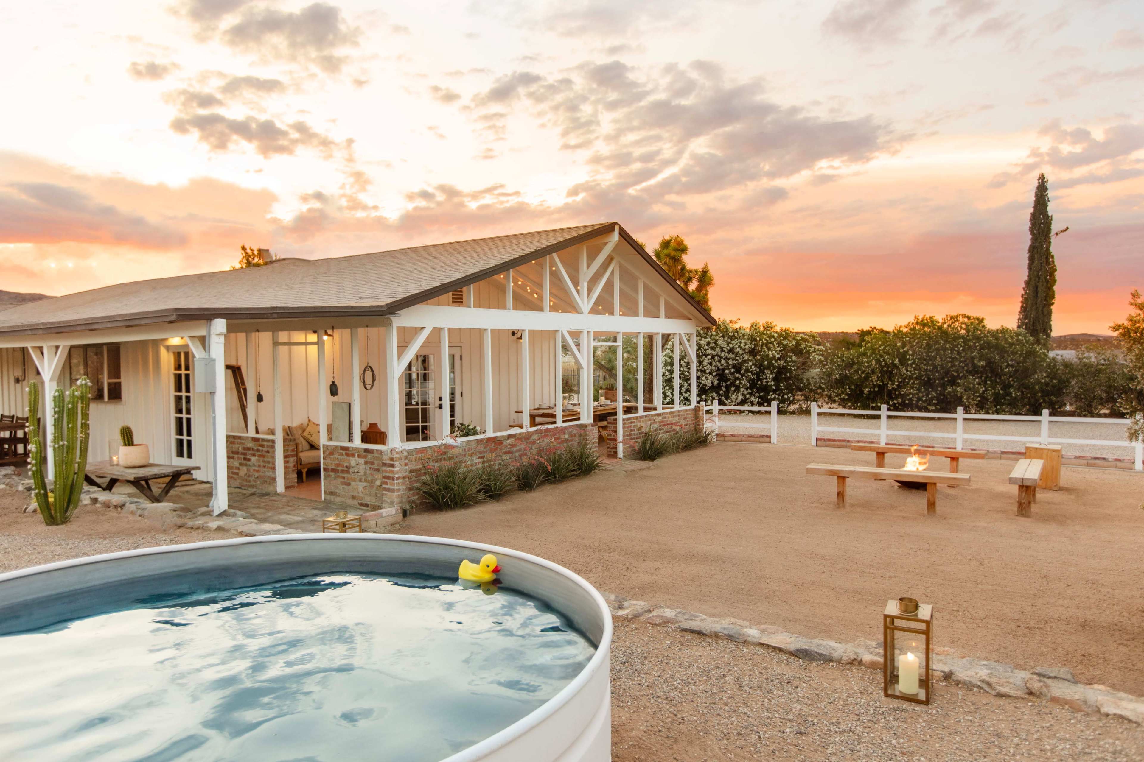 A single-story house with large windows and a pool sits in a desert landscape beneath a colorful sunset, accompanied by a fire pit and outdoor seating area.