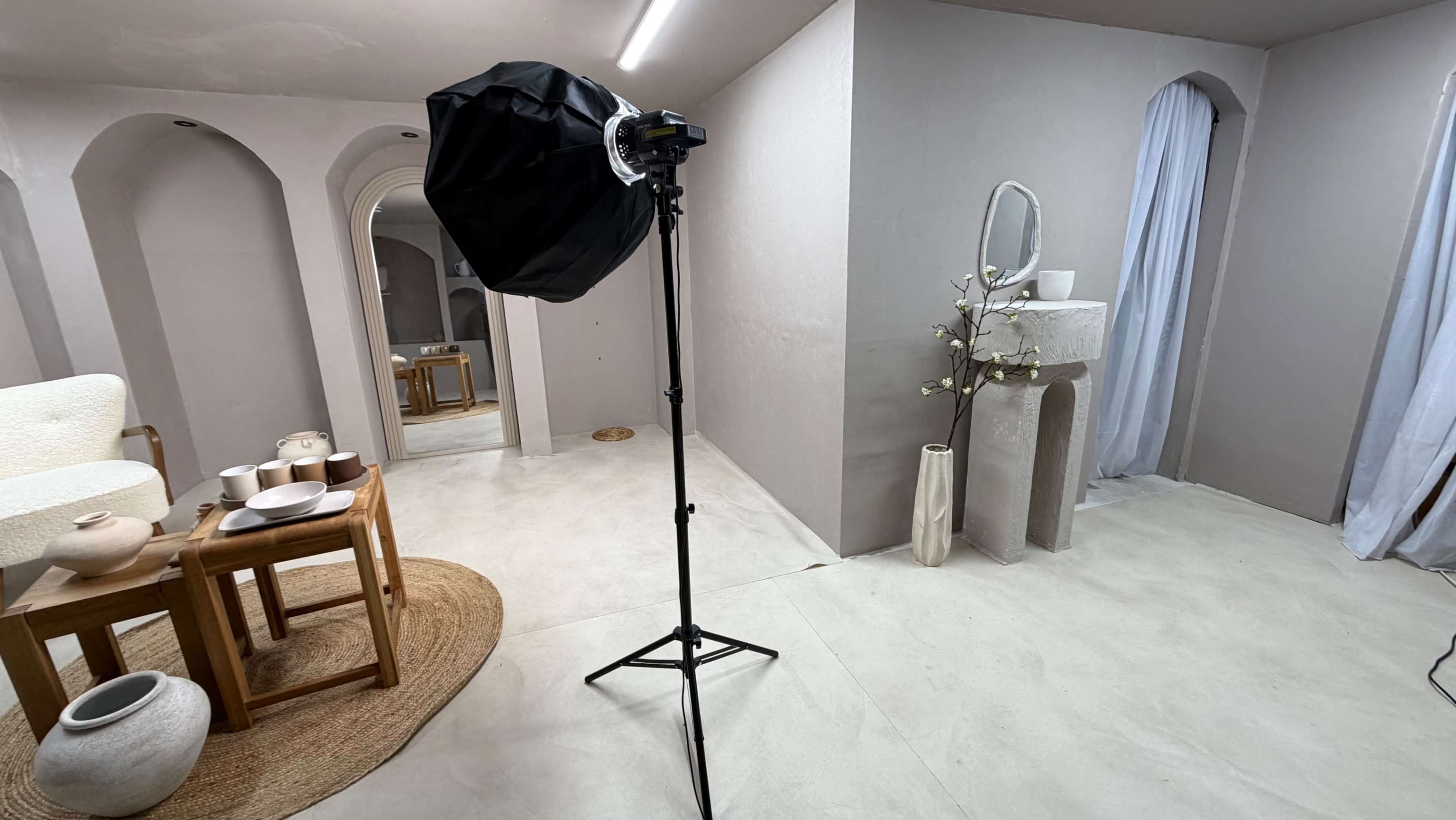 The image shows a minimalistic indoor setting with a lighting setup, a wooden table displaying pottery, and a mirror reflecting the space.