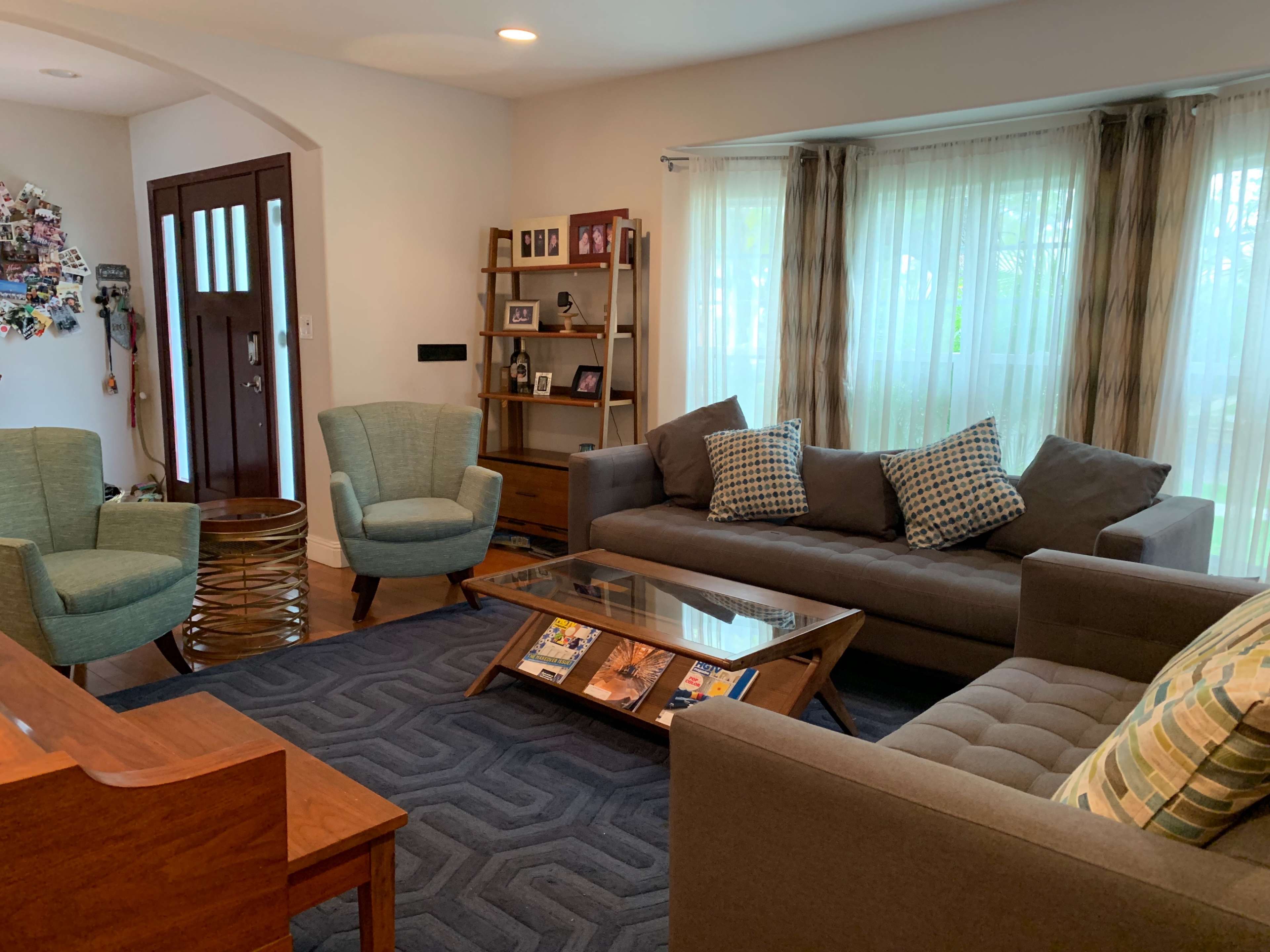 A cozy living room with two armchairs, a sofa, a wooden coffee table, and a bookshelf against a wall, all arranged around a patterned area rug and illuminated by natural light from large windows.
