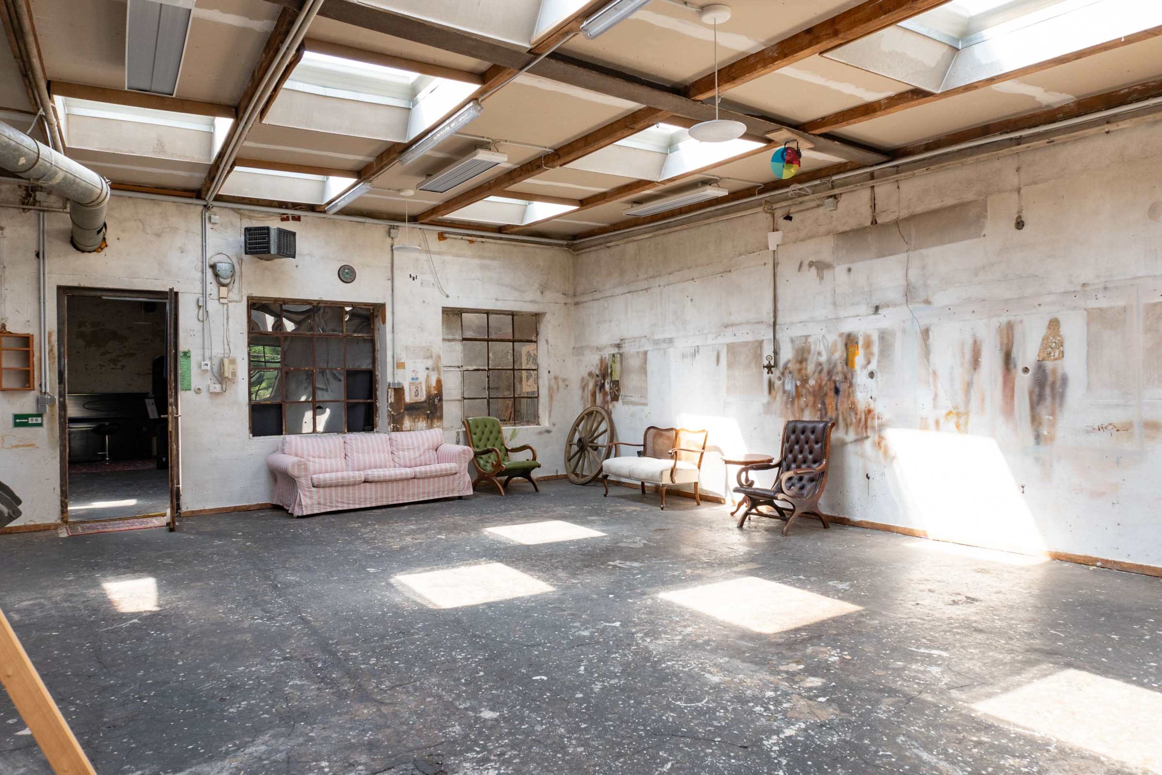 The image shows an empty industrial space with a wooden ceiling and several mismatched chairs and a couch arranged in the room.