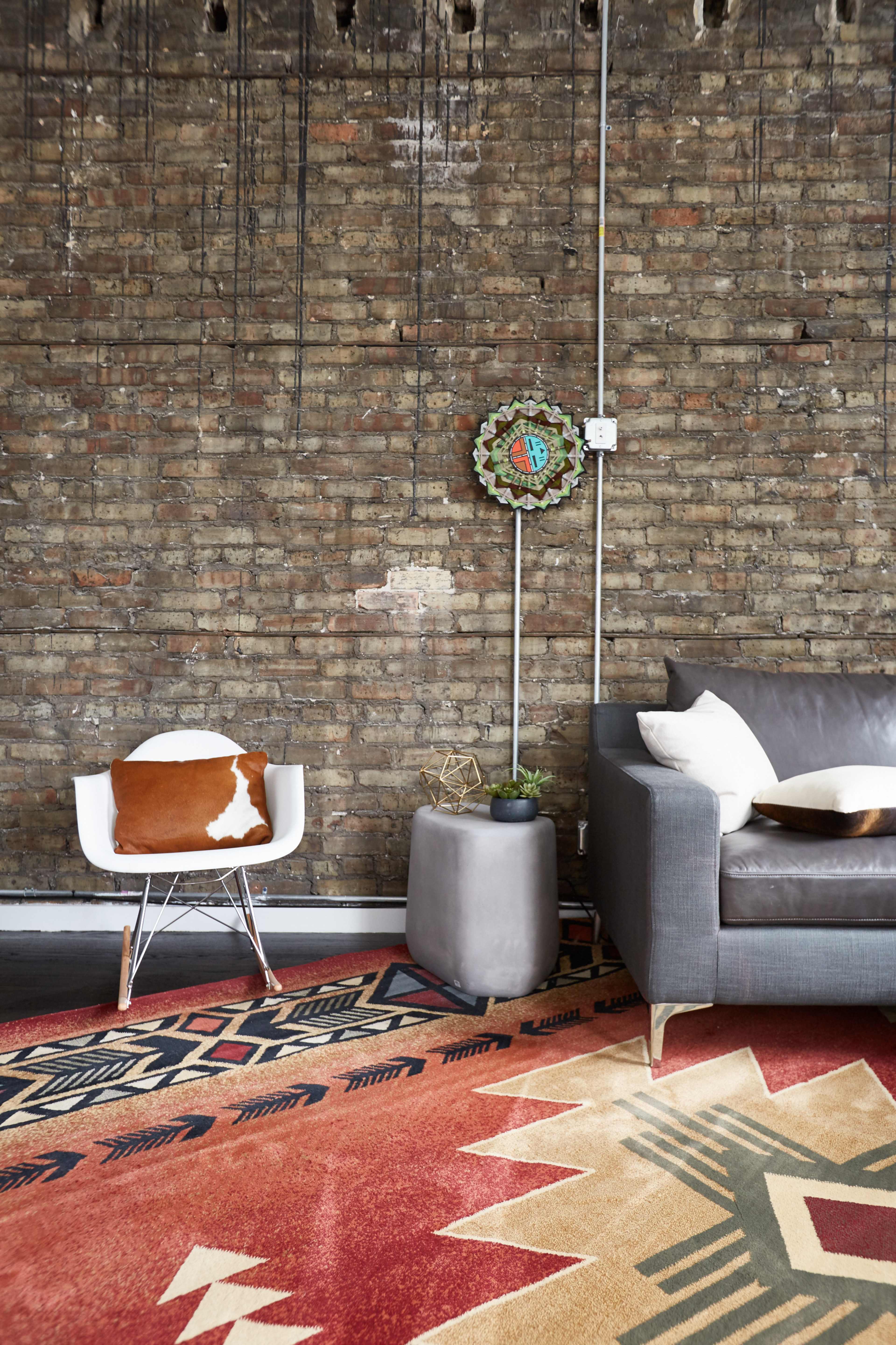 A modern living space featuring a gray sofa, a white chair with a decorative pillow, a small gray side table, a patterned rug, and a brick wall with an ornamental wall piece.