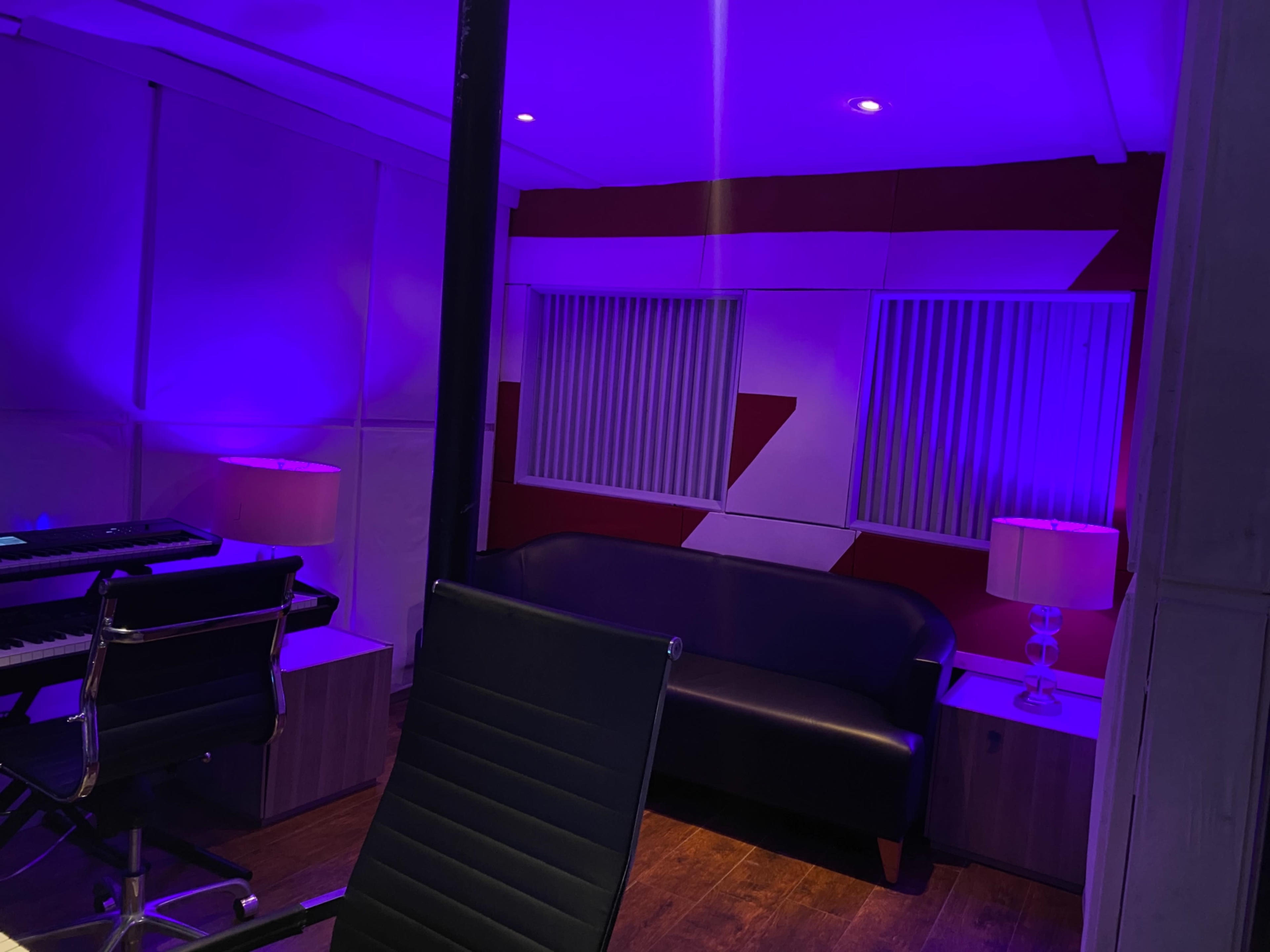 The image shows a modern studio room with purple lighting, a black sofa, a keyboard, and two lamps.