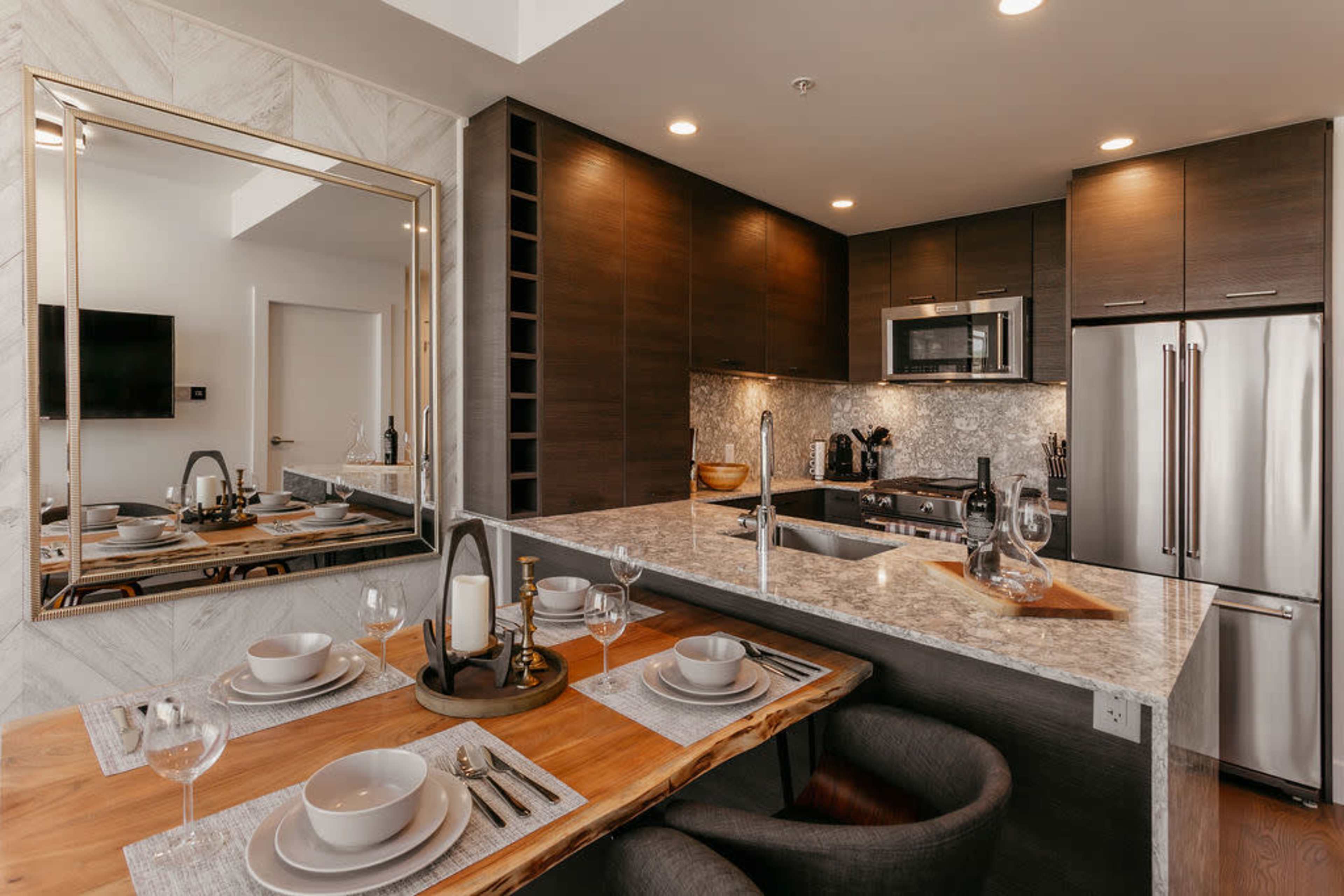 A modern kitchen and dining area featuring a marble countertop, dark cabinetry, and a large mirror reflecting the dining table set with plates and glasses.