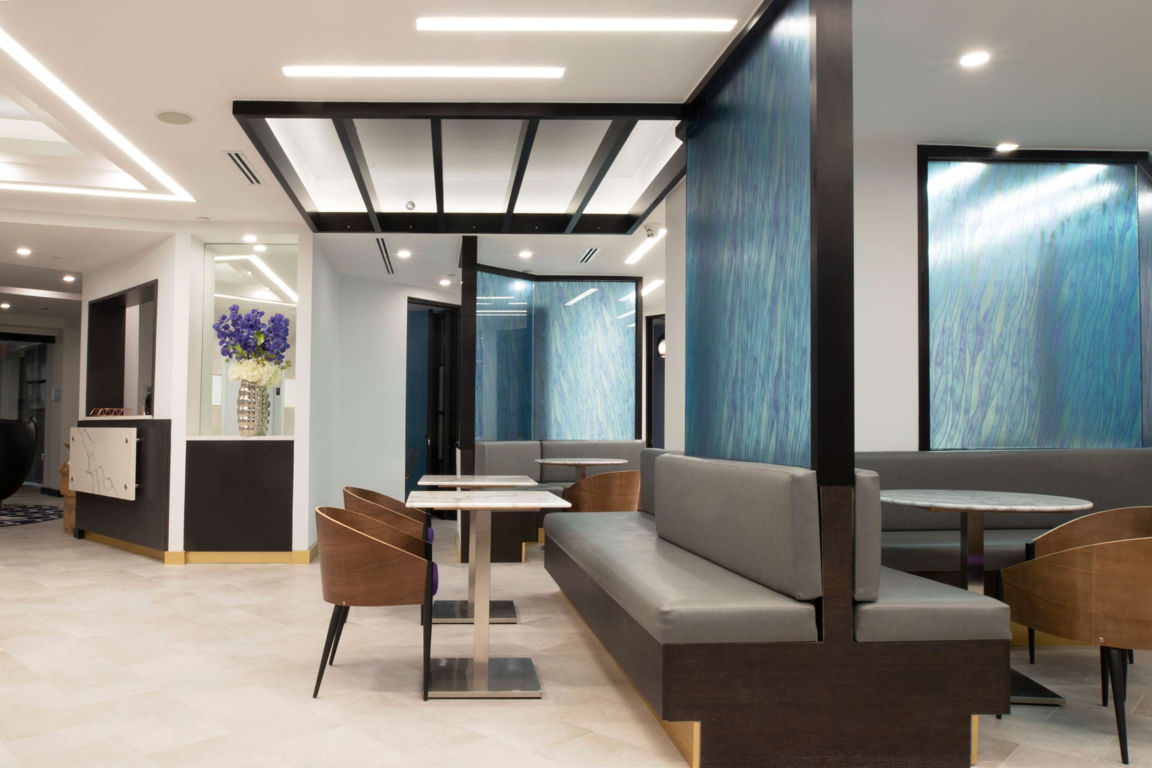 A modern interior space featuring a seating area with gray benches and wooden chairs, complemented by blue accent walls and decorative floral arrangements.