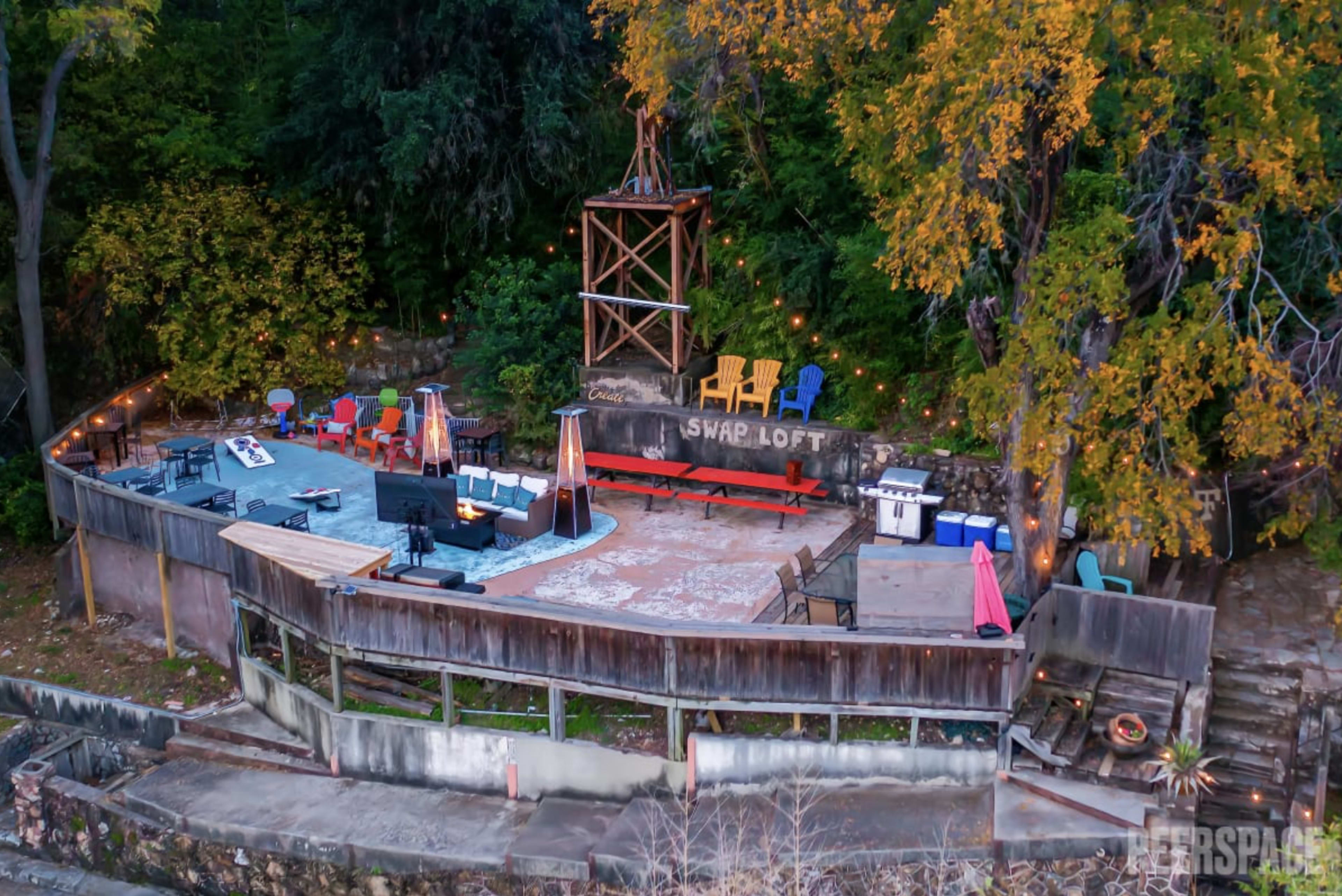 An outdoor gathering area with a stage, seating options, and decorative lighting surrounded by trees.