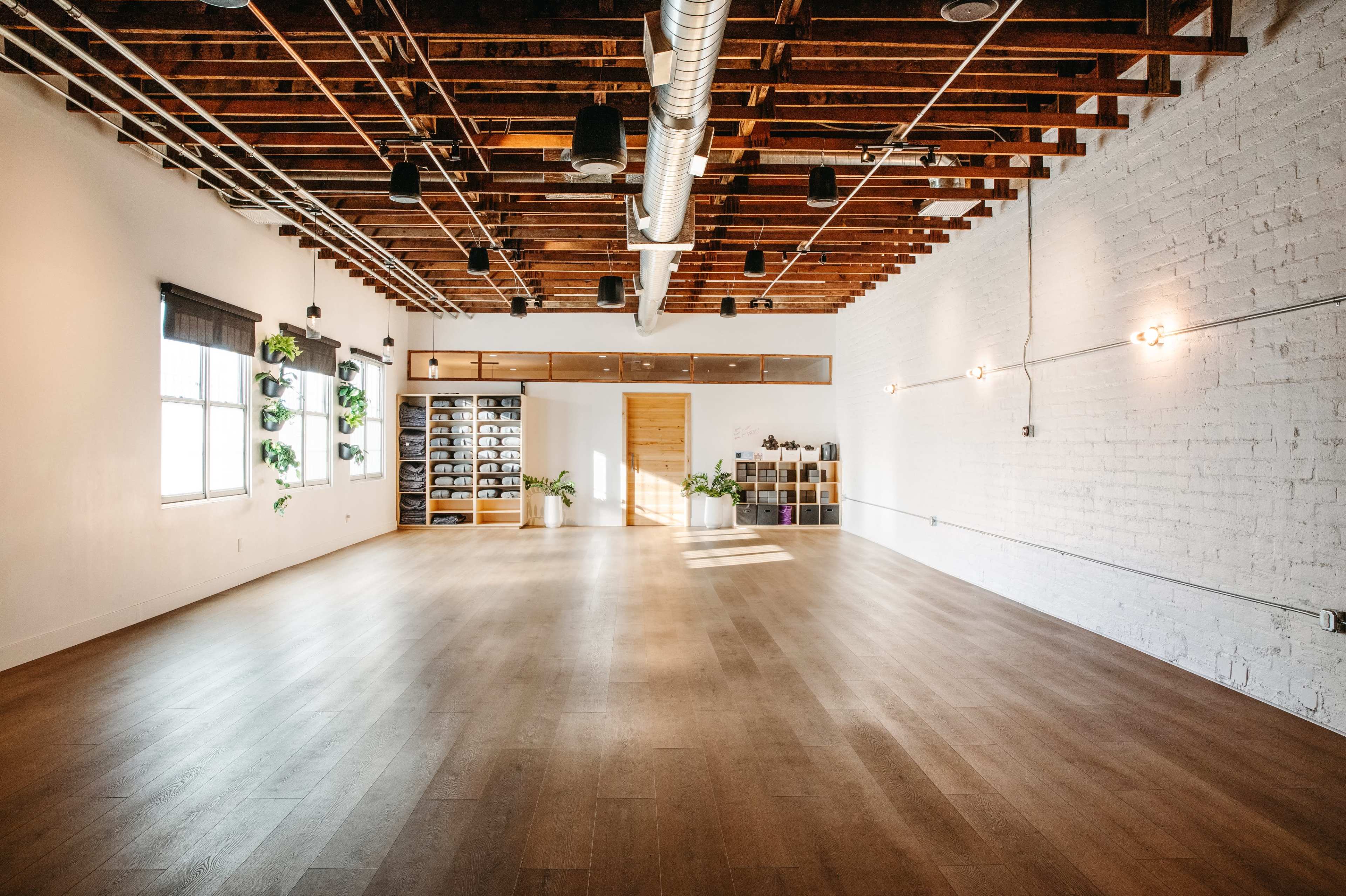 The image shows a spacious, well-lit studio with wooden flooring, exposed wooden beams, and minimalistic decor, featuring plants and shelving against the walls.