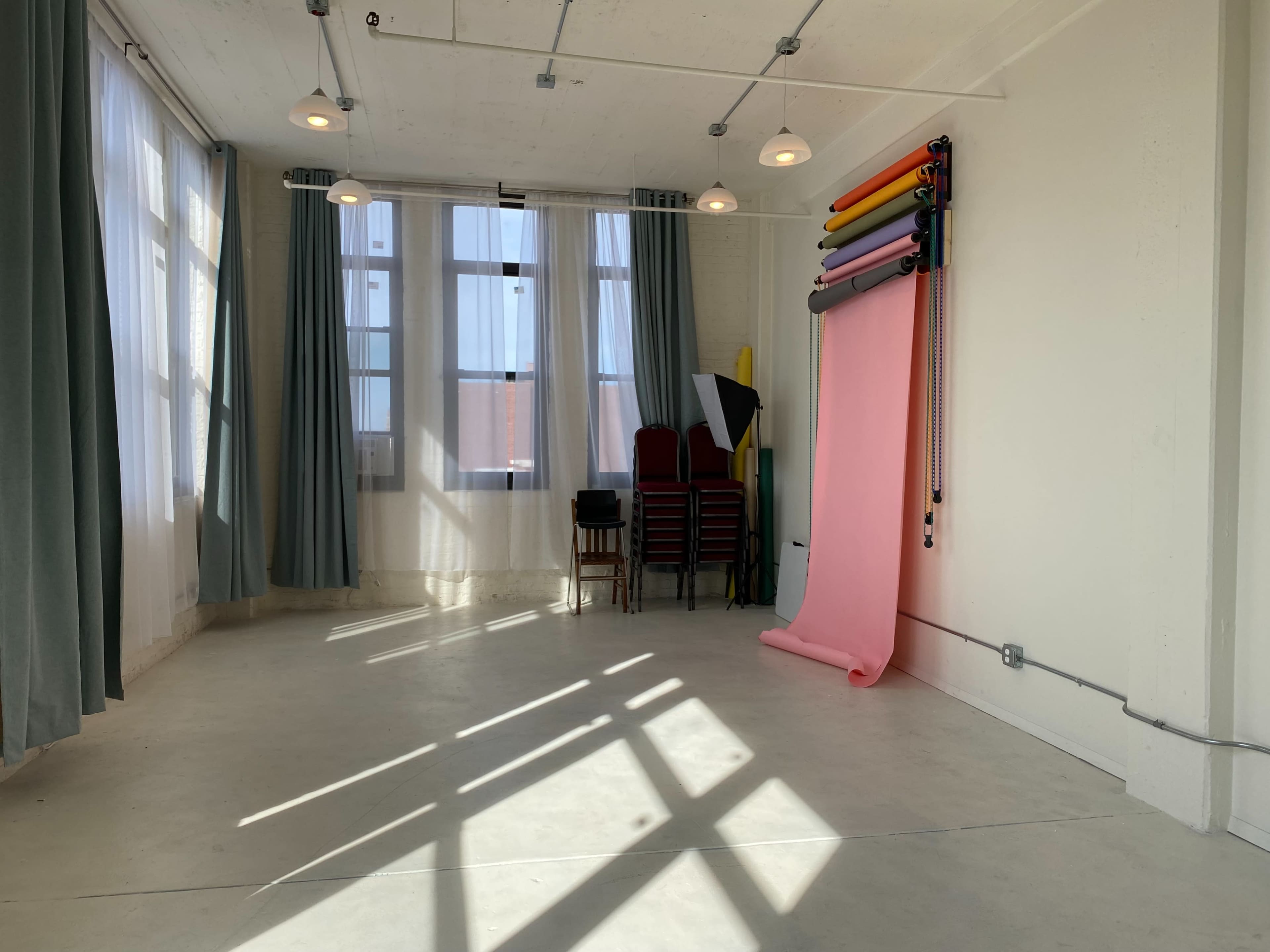A bright room with large windows, green curtains, a stack of chairs, and a roll of photography backdrop paper in various colors.