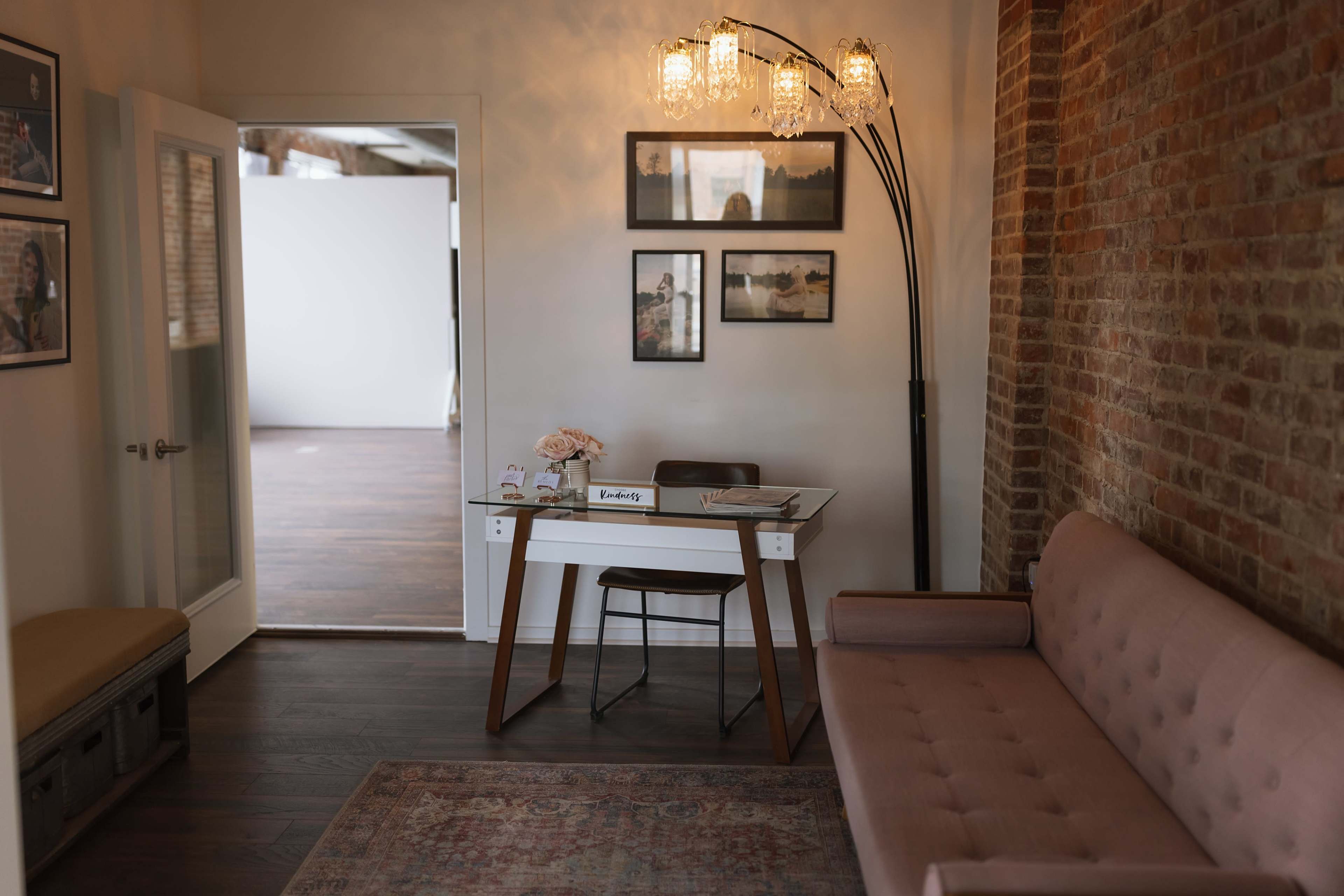 A modern workspace features a pink sofa, a wooden desk with a floral arrangement, and framed artwork on the wall.