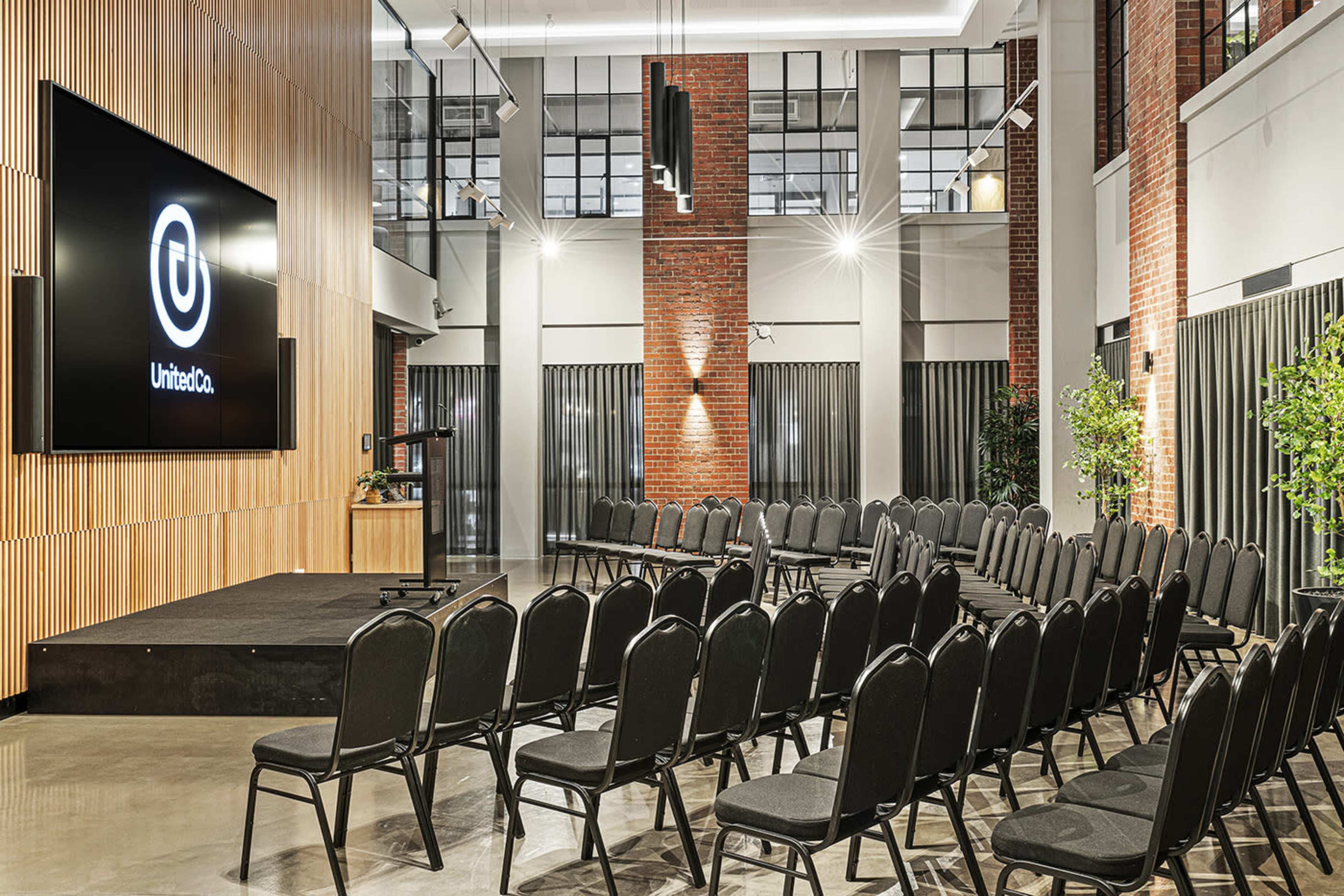 The image shows a spacious event room with rows of black chairs facing a large screen and a wooden wall, illuminated by overhead lights and featuring brick columns.