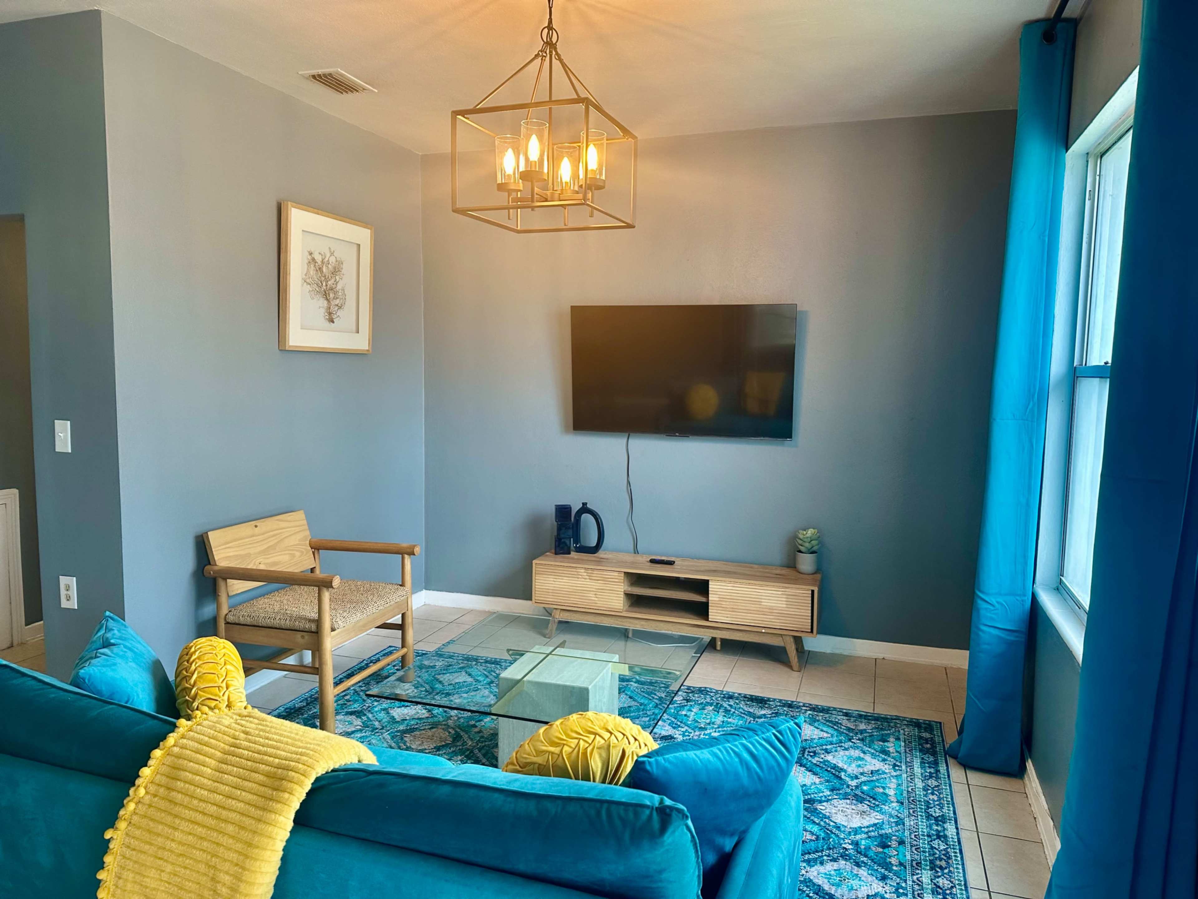 The living room features a teal sofa, a wooden chair, and a mounted television against a gray wall, with a blue area rug under a glass coffee table.