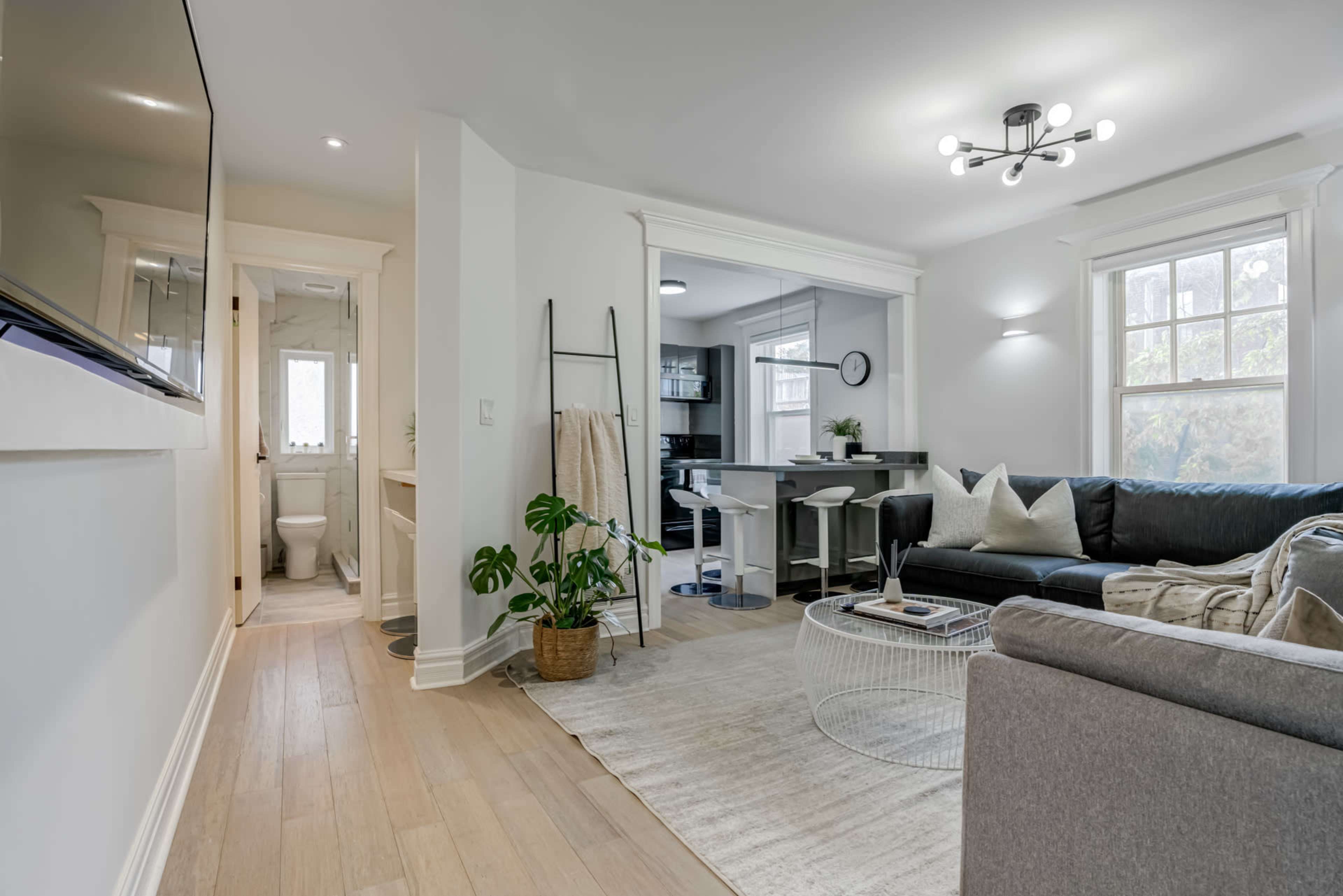 A modern, open-concept living space with a dark sofa, light hardwood floors, and a visible kitchen area.