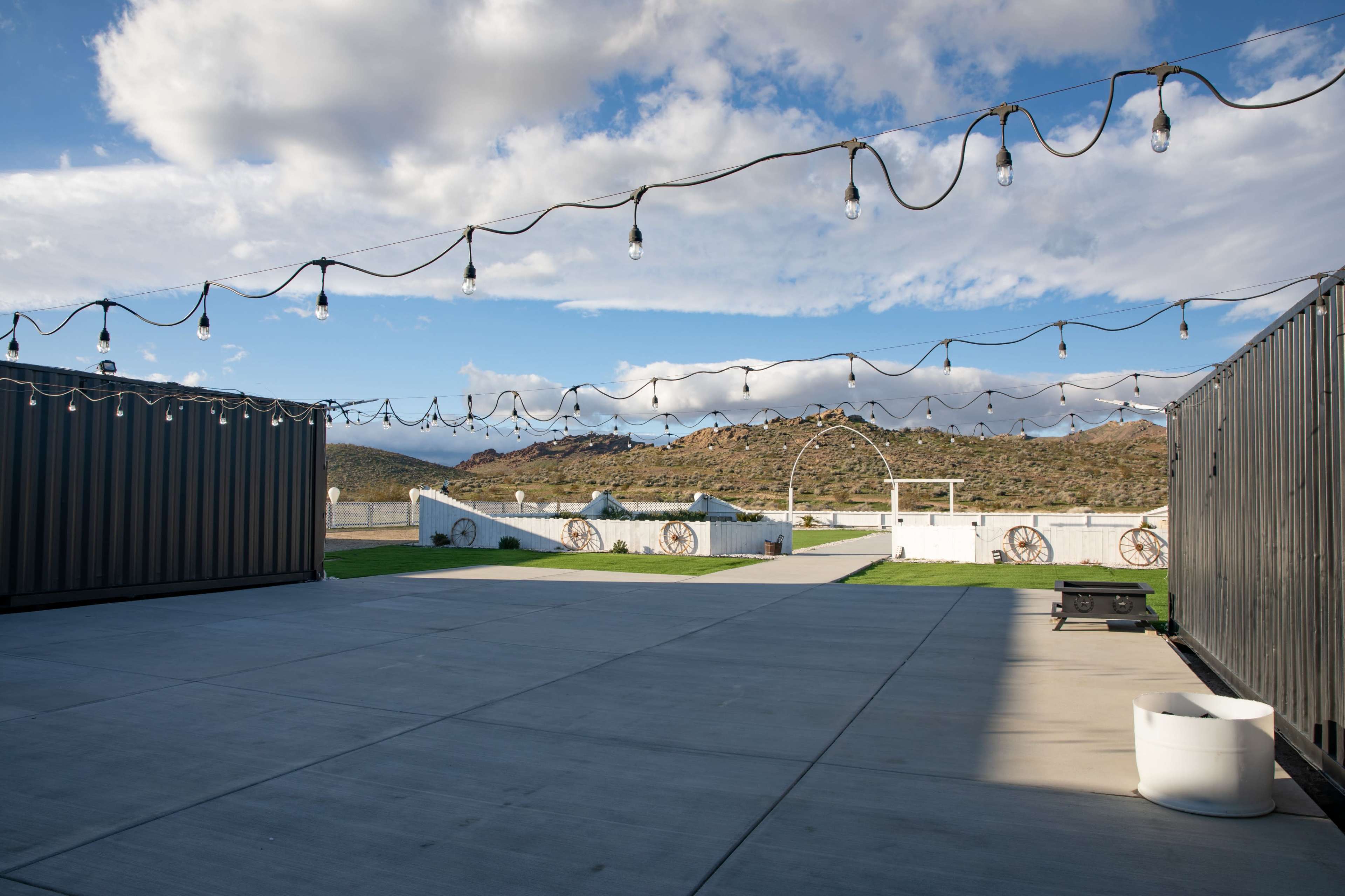 The scene shows a spacious outdoor area with string lights overhead, surrounded by shipping containers and with a view of mountains in the background.