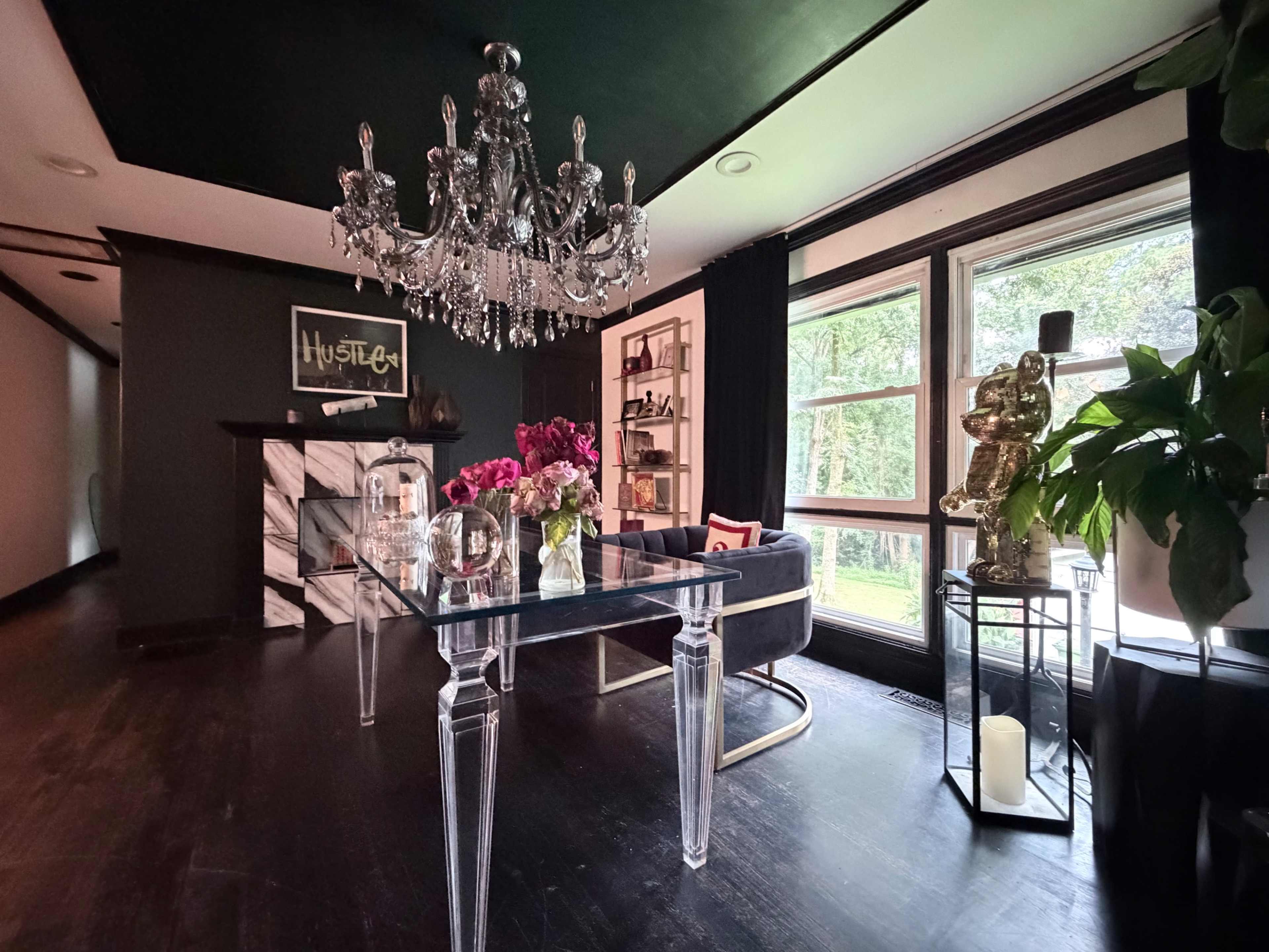 The image shows a modern room featuring a glass table, a chandelier, a marble fireplace, and large windows with a view of greenery outside.
