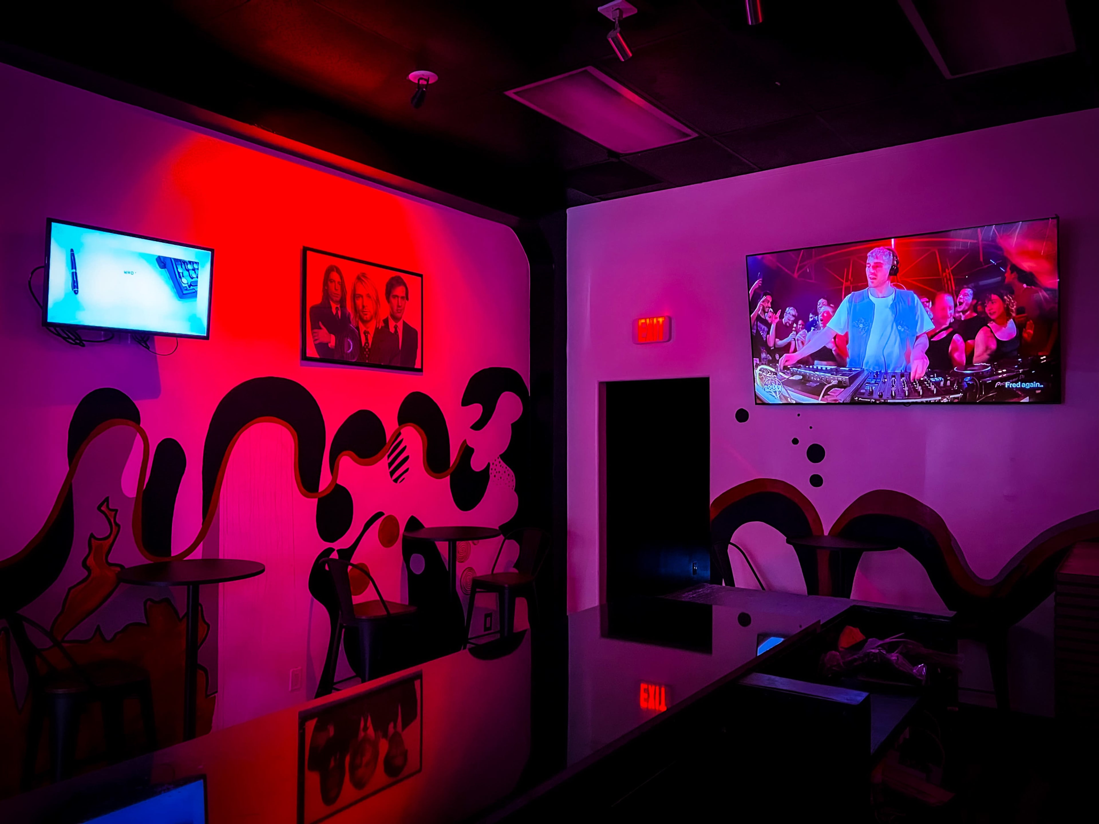 The interior of a dimly lit bar features a mural on the wall, two televisions displaying different images, and tables.