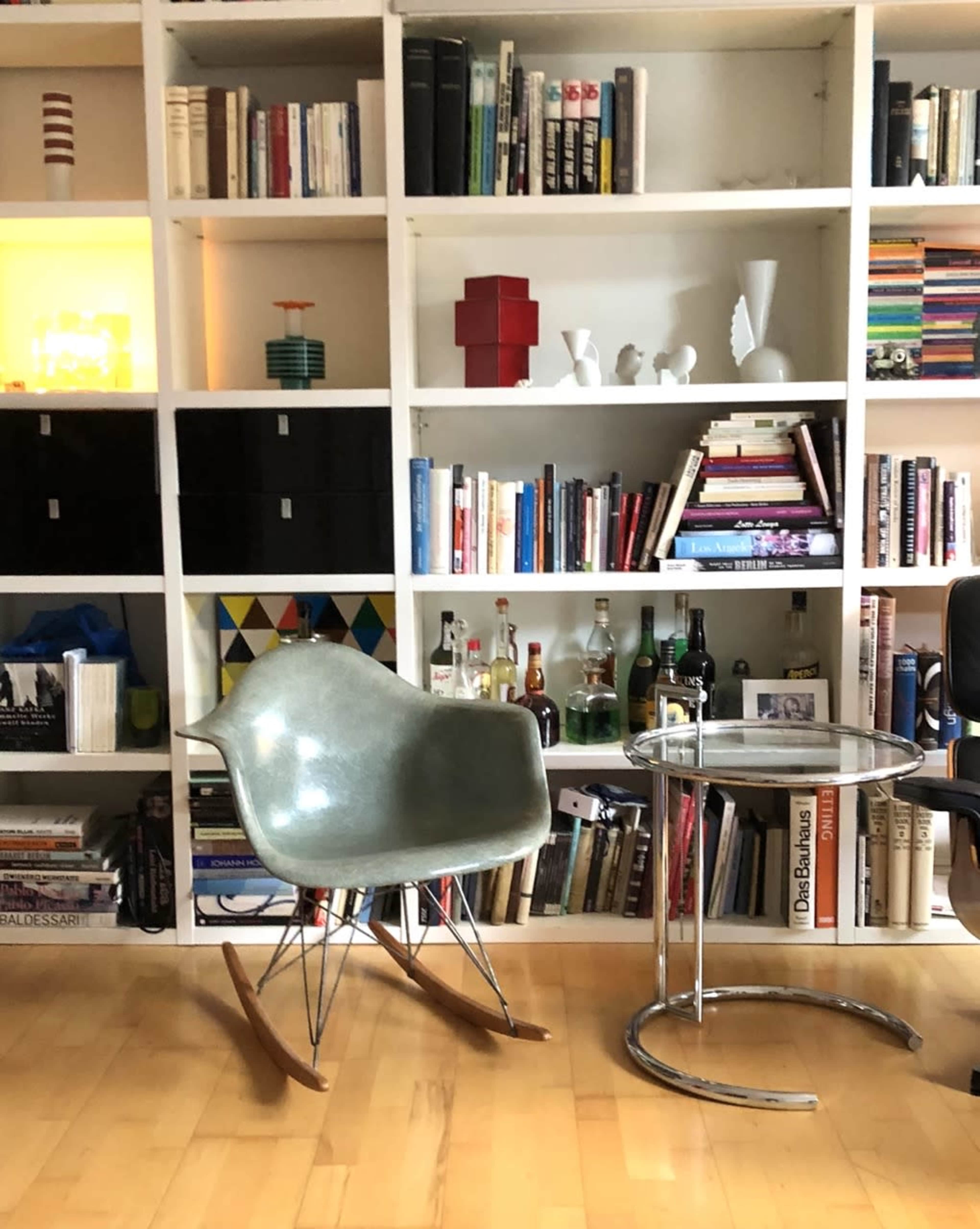 A curved, light green rocking chair sits beside a small glass side table in front of a white bookshelf filled with various books and decorative items.