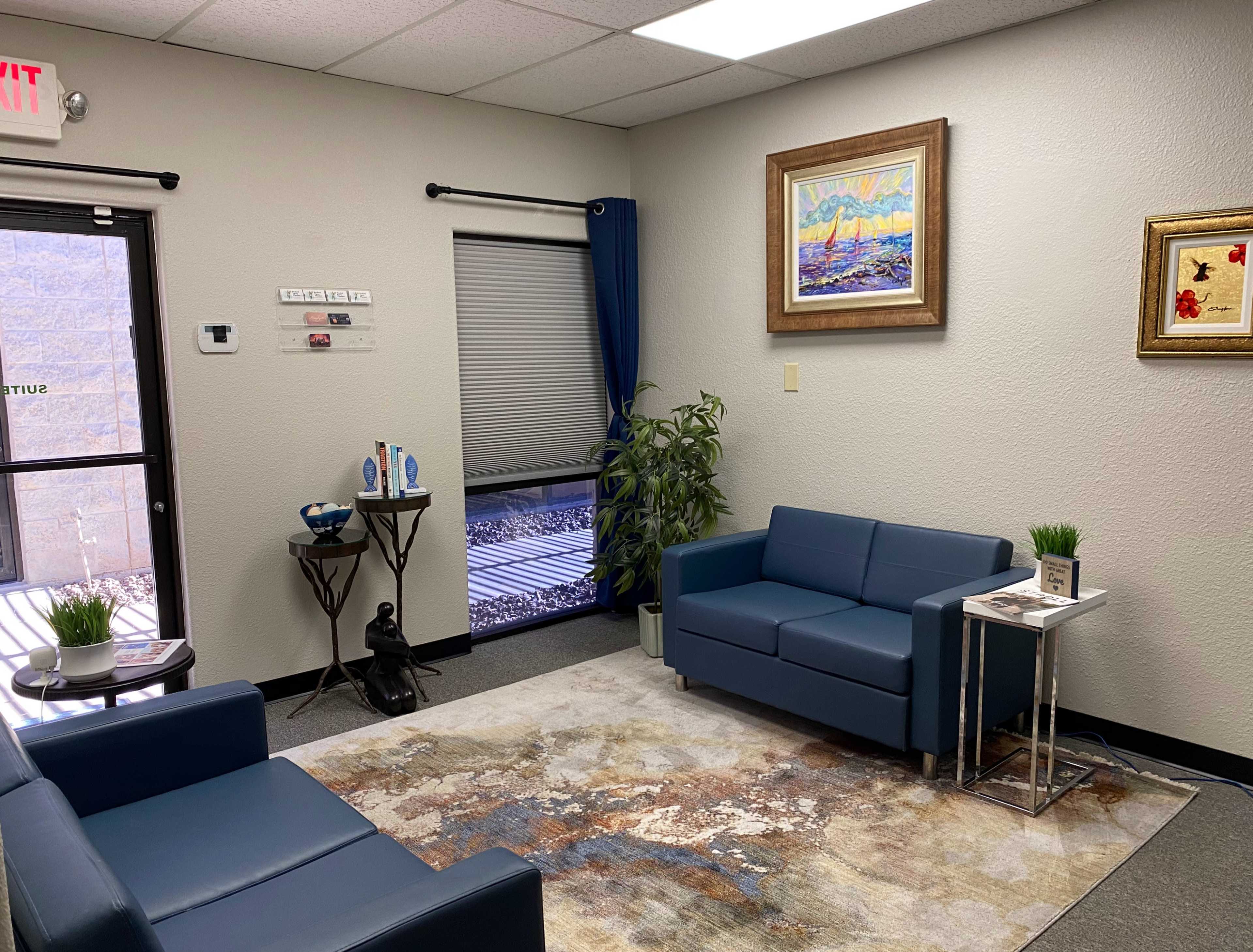 The image shows a waiting room with two blue couches, a small table, a decorative plant, and artwork hanging on the walls.