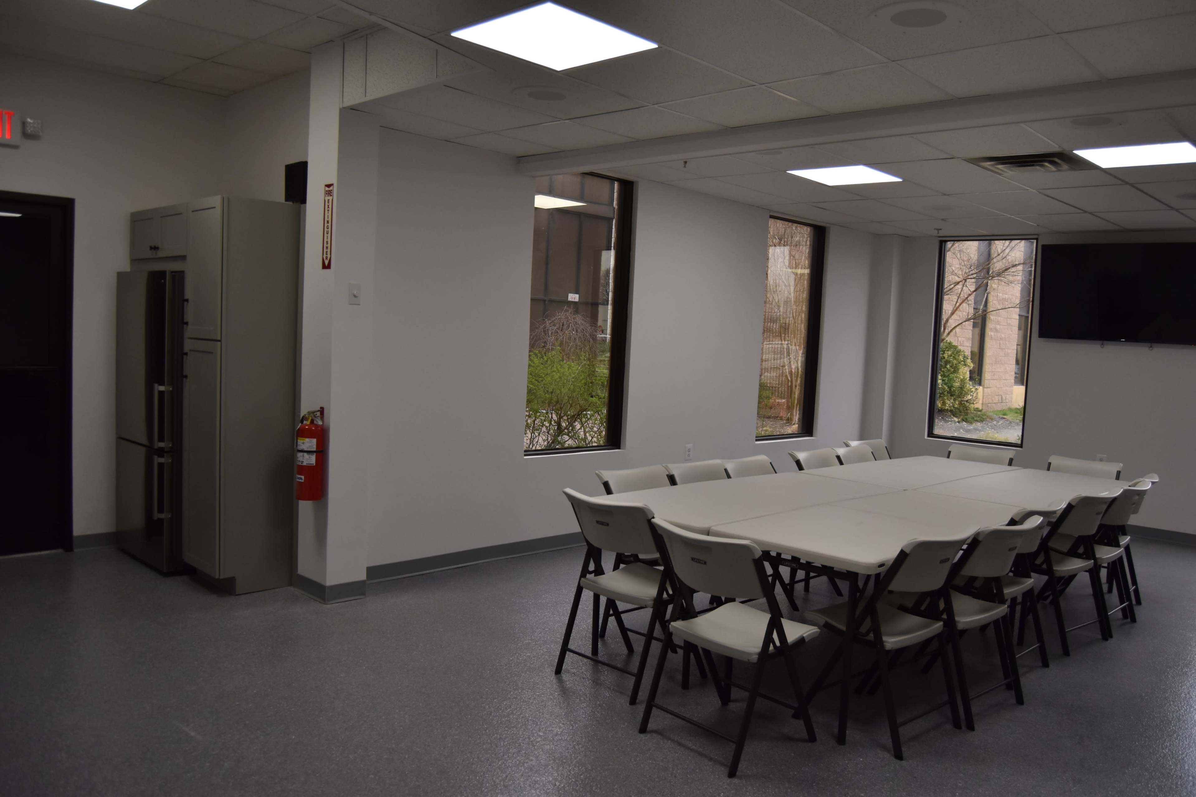 A meeting room with a rectangular table surrounded by chairs, a refrigerator in the corner, and large windows letting in natural light.