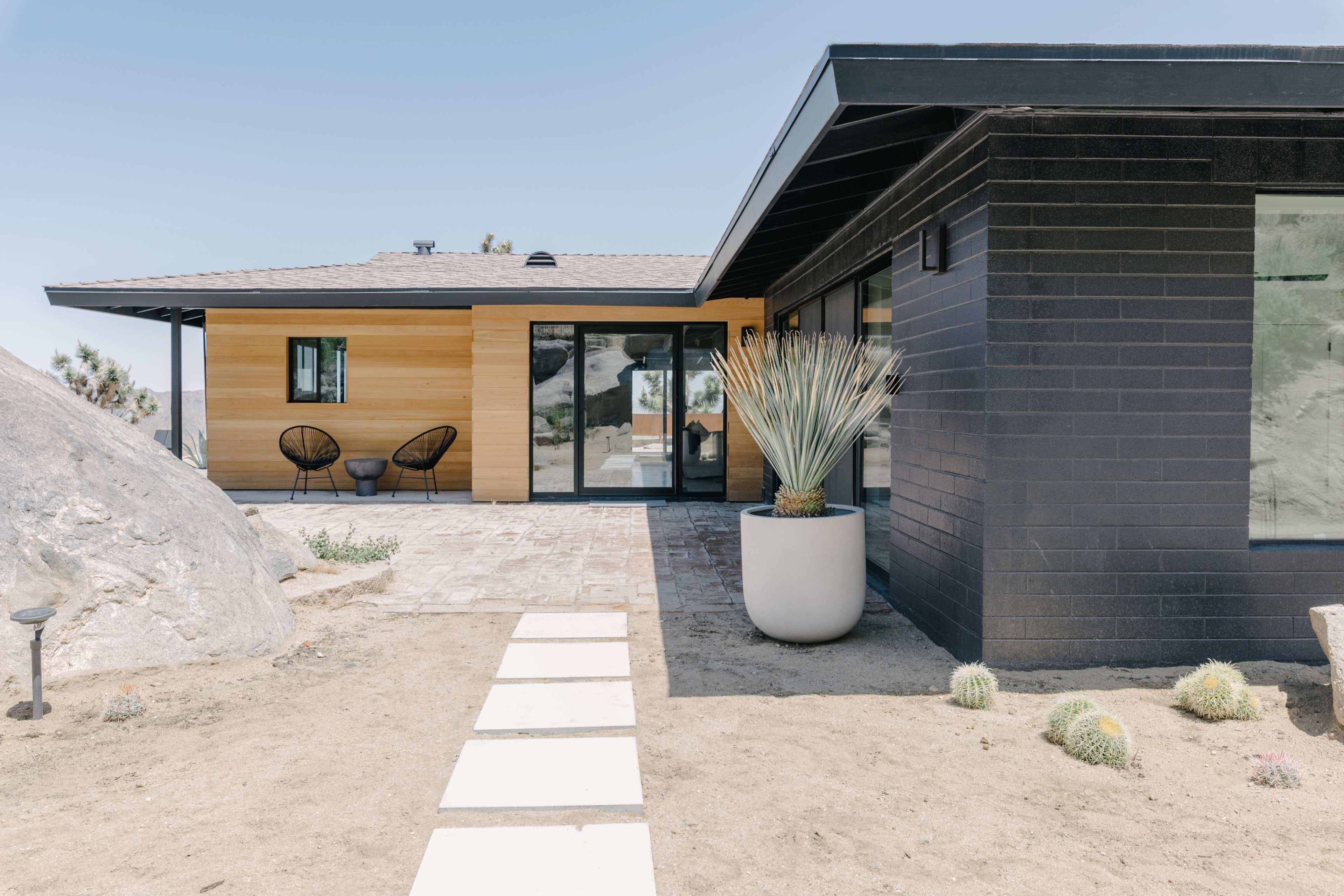 A modern house with a combination of wood and black brick exteriors, featuring a landscaped yard with a stone pathway and a large potted plant.