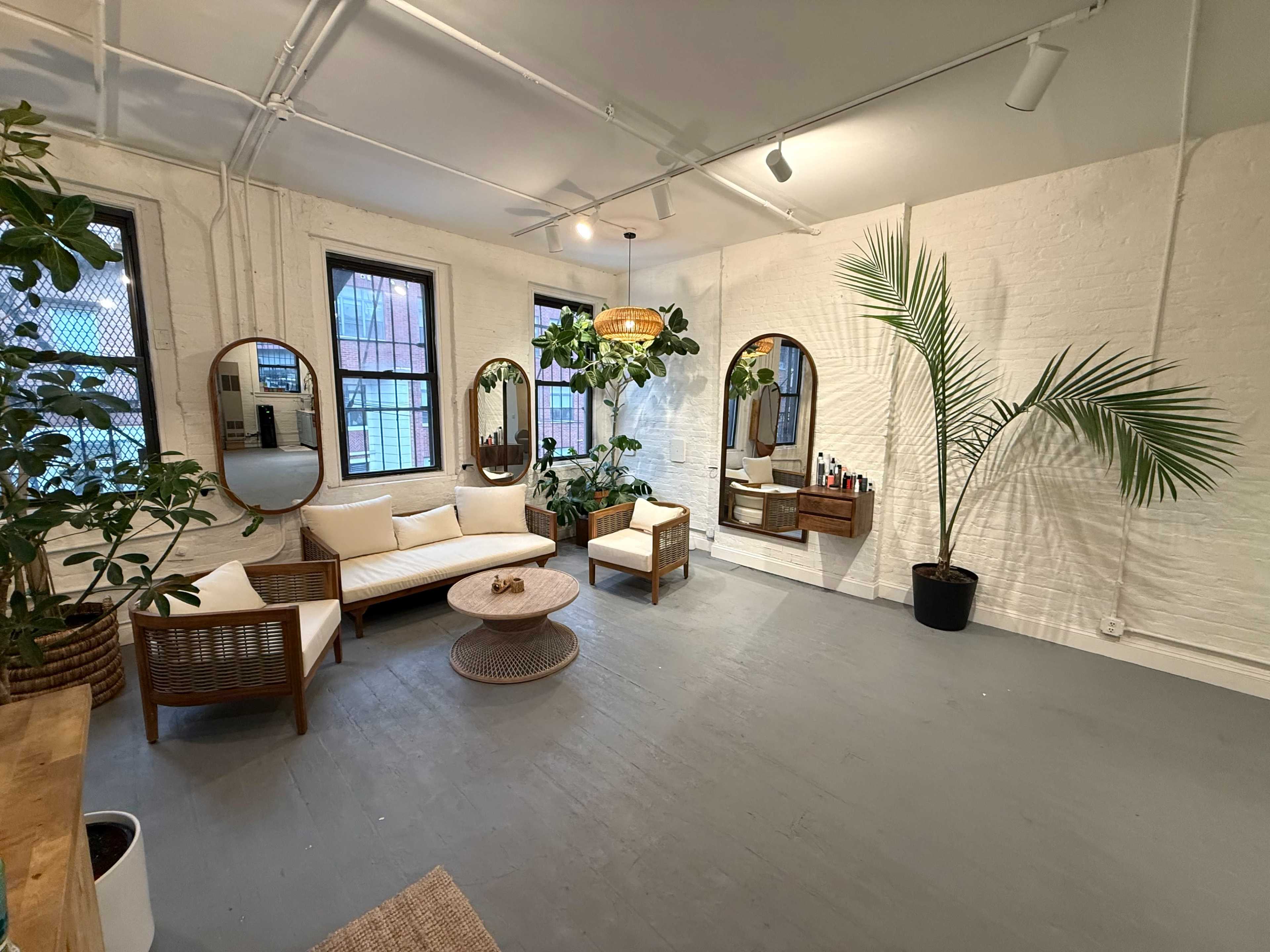 The image shows a bright, open room furnished with a light-colored sofa, a circular coffee table, and several plants, along with two mirrors on the walls and a small cabinet displaying various items.