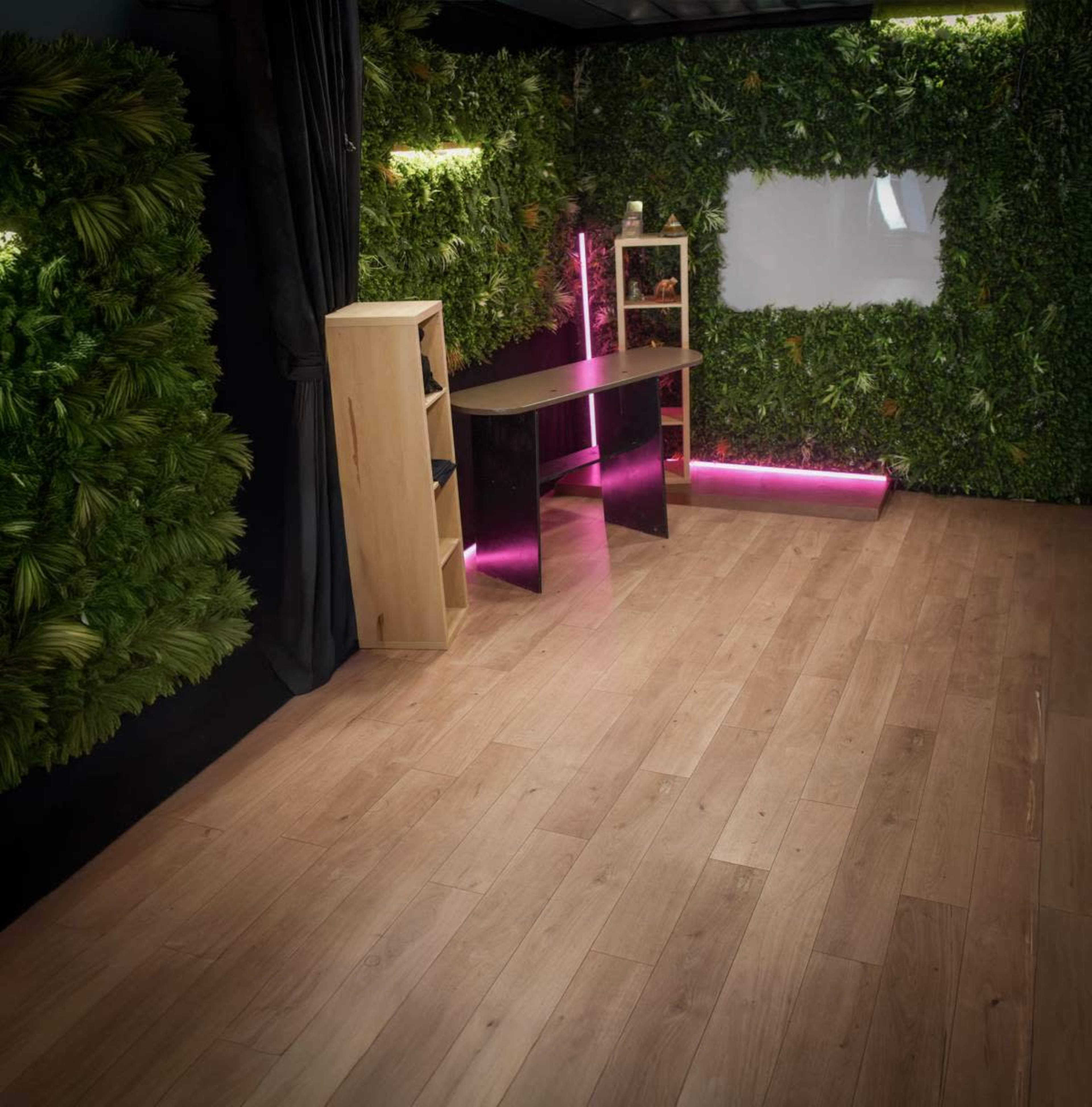 The image shows a modern room with wooden flooring, a sleek black table, and walls adorned with lush green foliage.