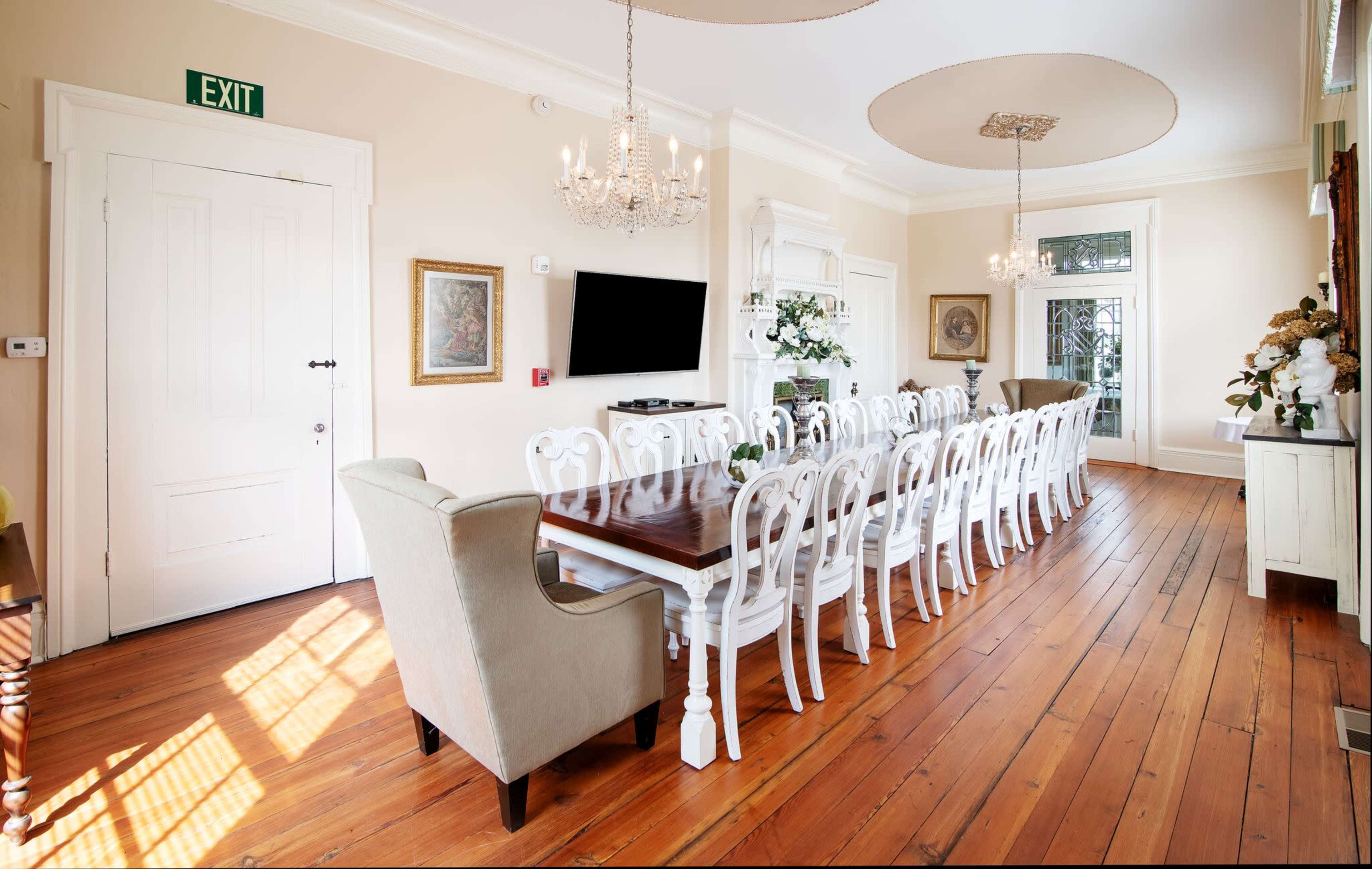 A spacious dining room features a long wooden table surrounded by white chairs, with a chandelier overhead and an exit sign on the wall.
