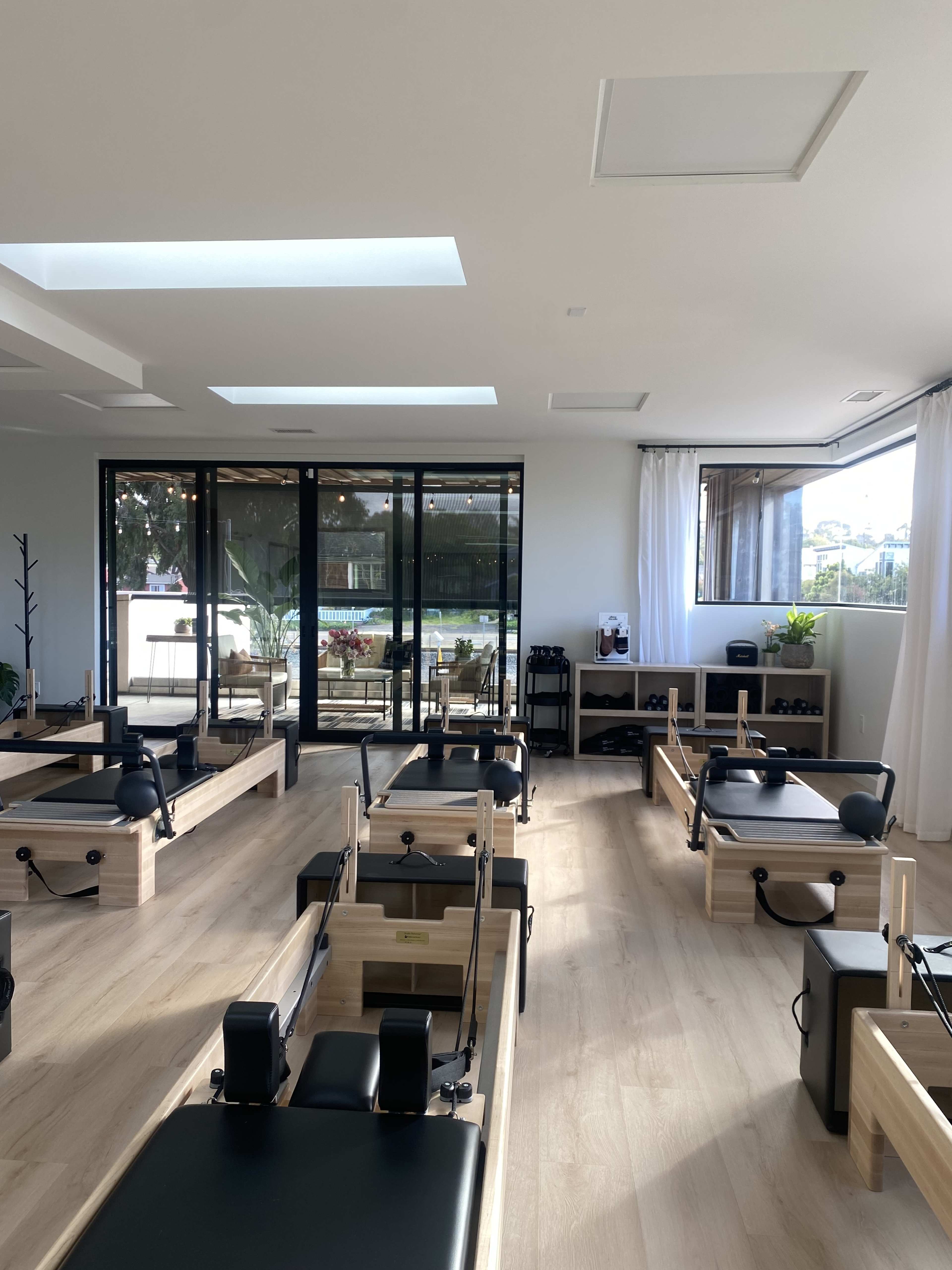 The image shows a modern Pilates studio with several reformer machines arranged neatly, large windows allowing natural light, and a view of an outdoor area.
