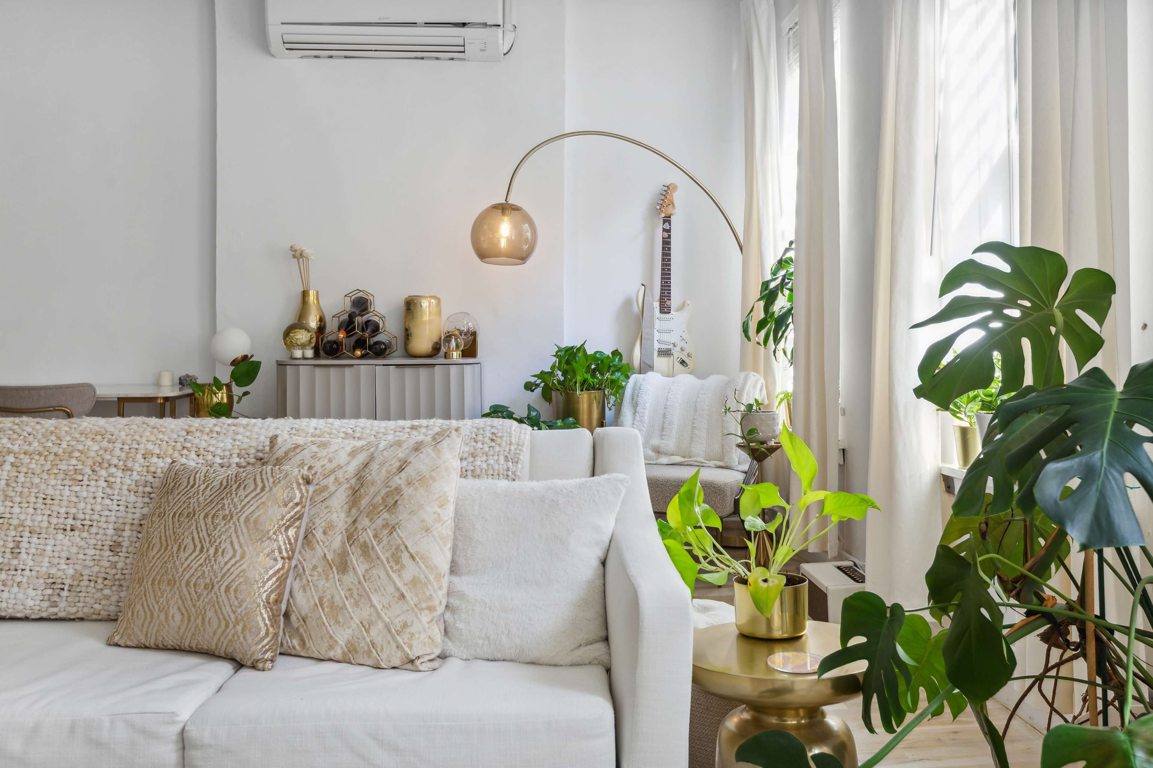 The image shows a stylish living room with a white sofa adorned with decorative pillows, plants, and a guitar positioned next to a minimalist side table.