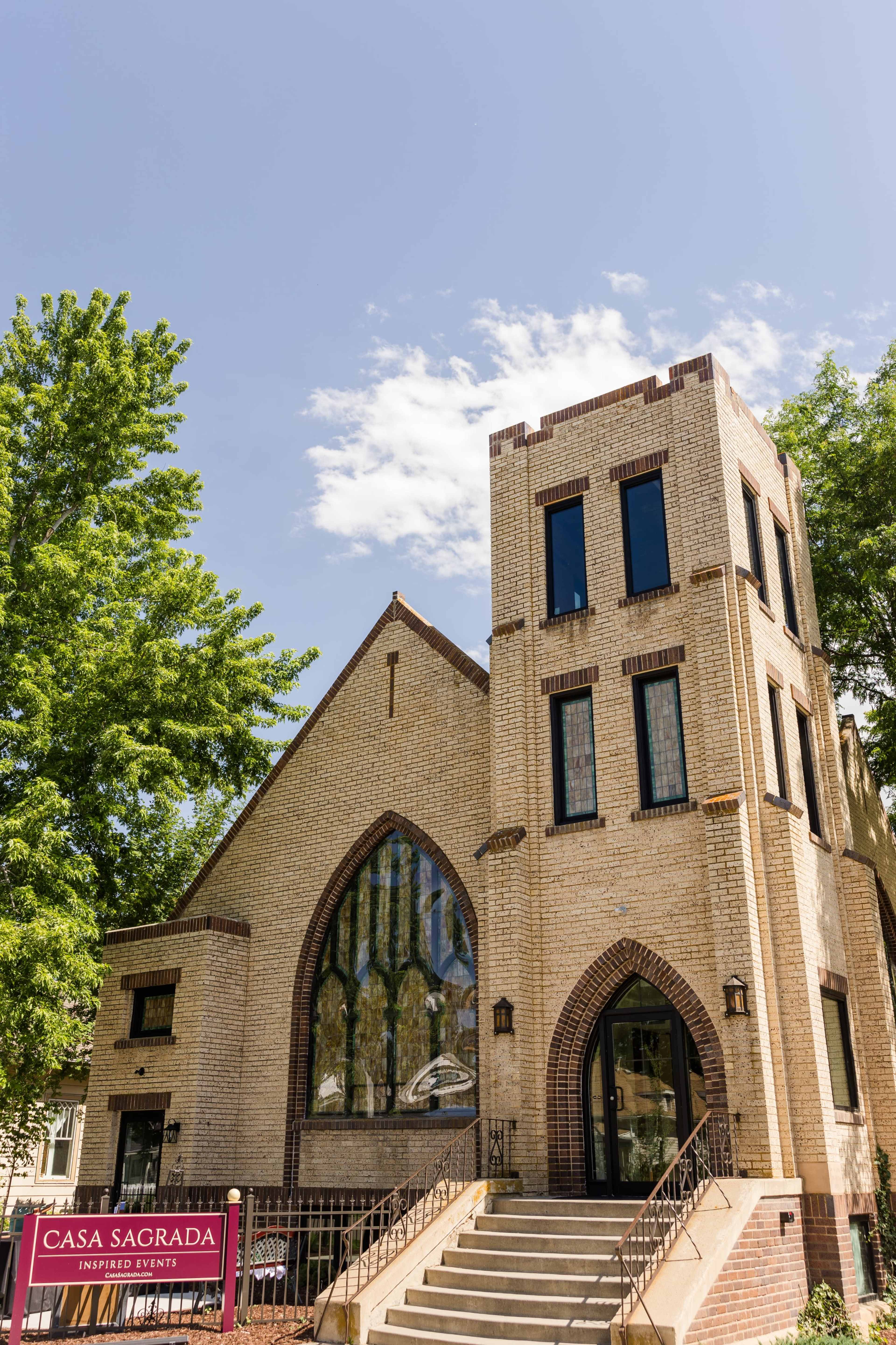 A light-colored brick building with large arched windows and a decorative entrance, accompanied by a sign reading "Casa Sagrada" for inspired events.