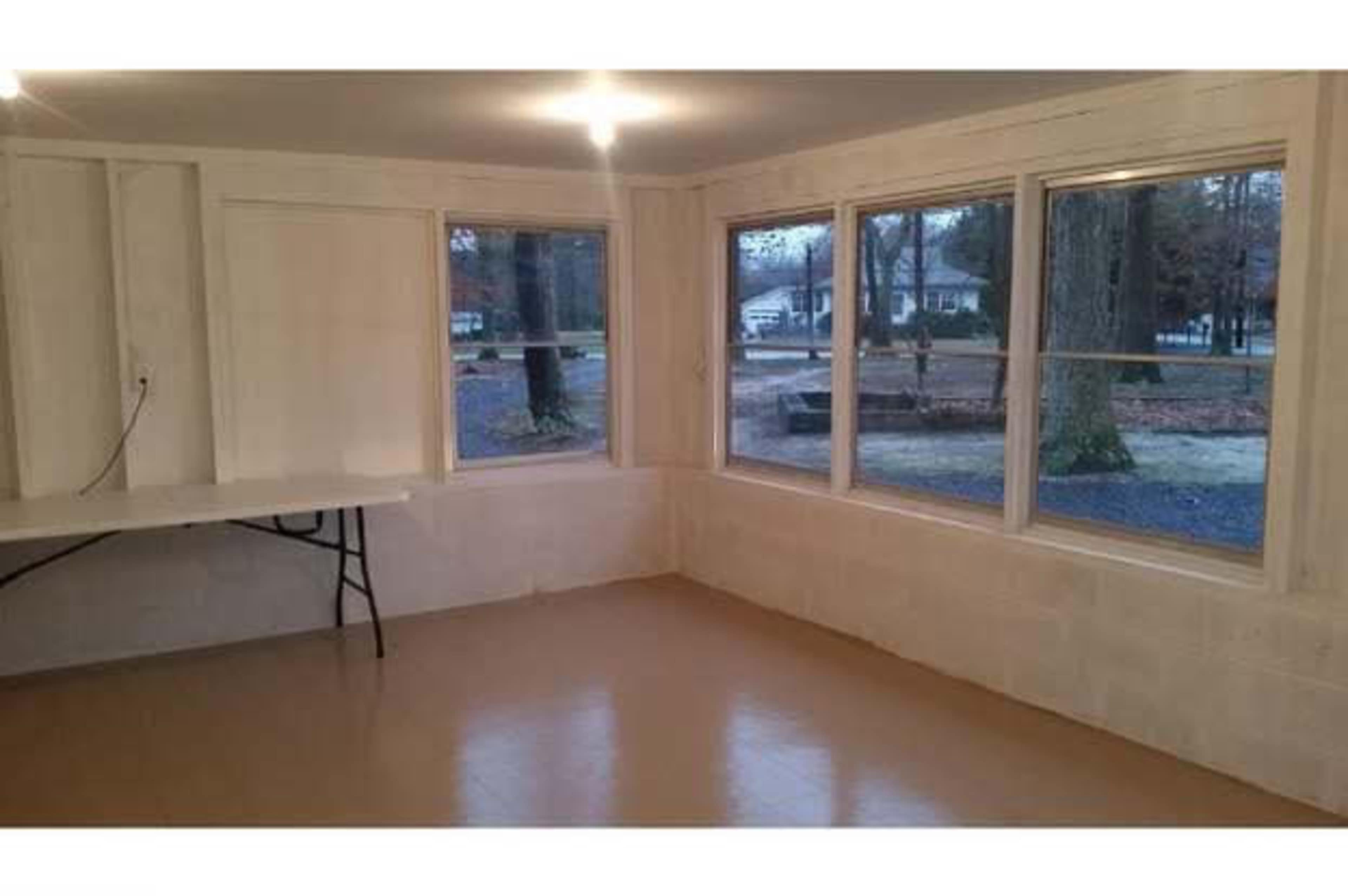 The image shows a vacant room with large windows, a folding table against one wall, and a smooth floor.