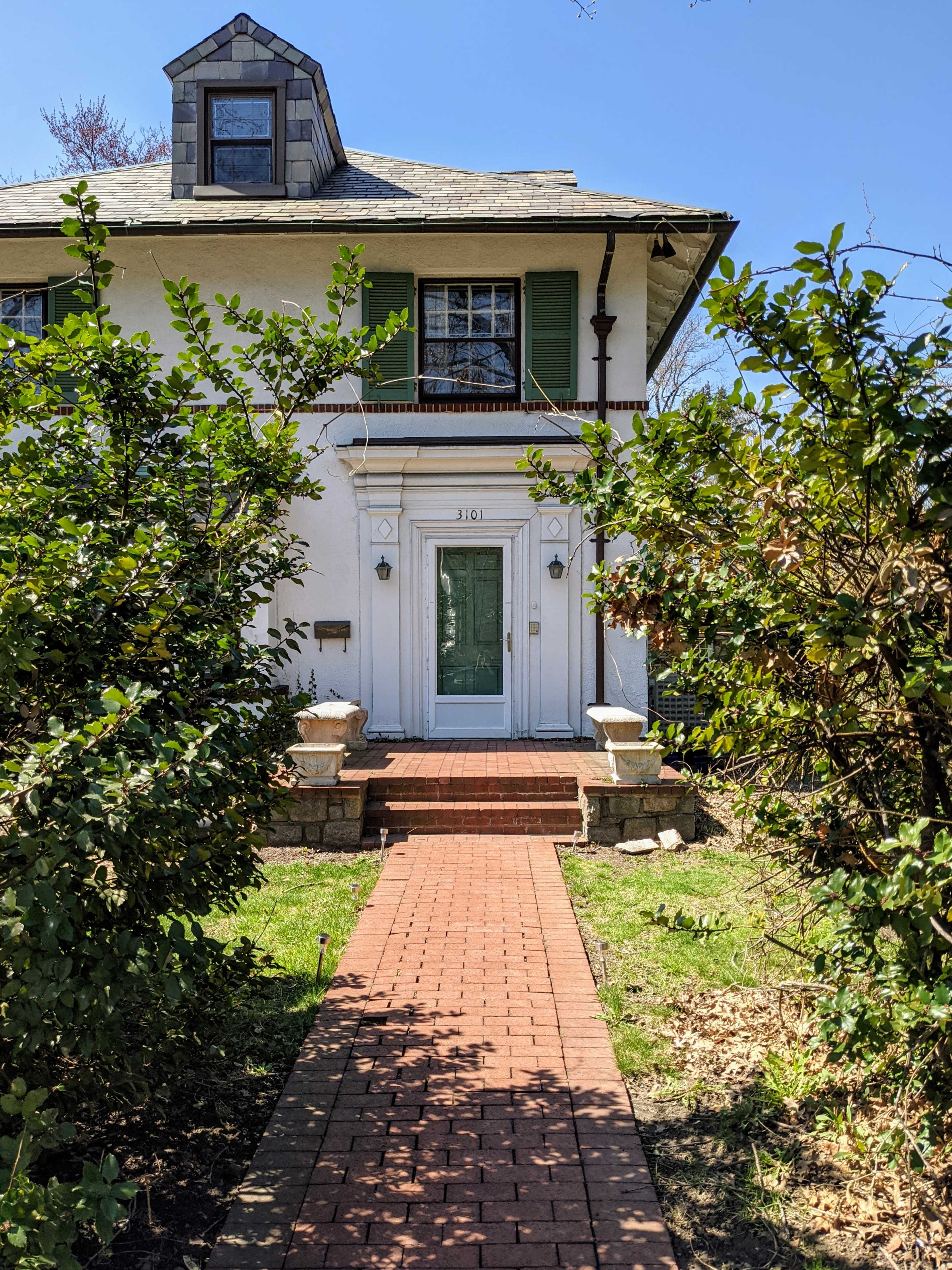 A brick path leads to the entrance of a two-story white house with green shutters and surrounding greenery.