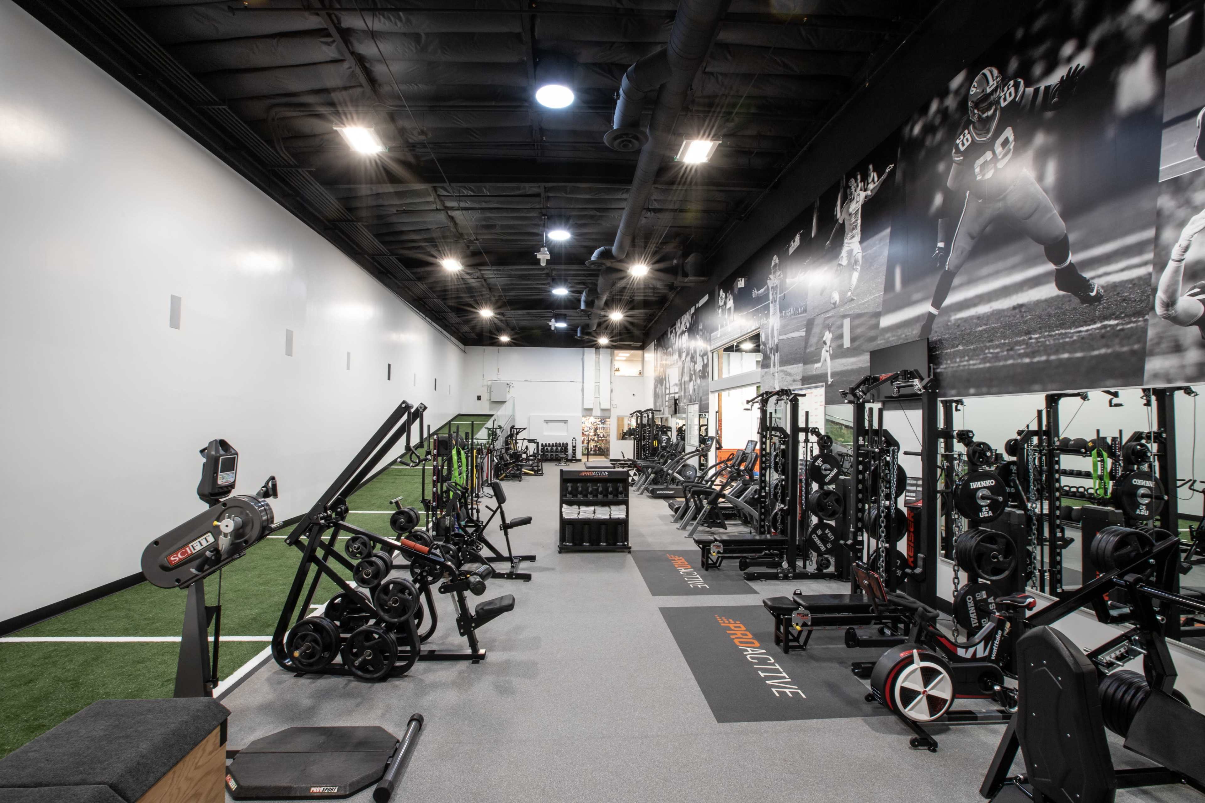 The image shows a spacious gym equipped with various fitness machines, free weights, and equipment, alongside an artificial turf area.