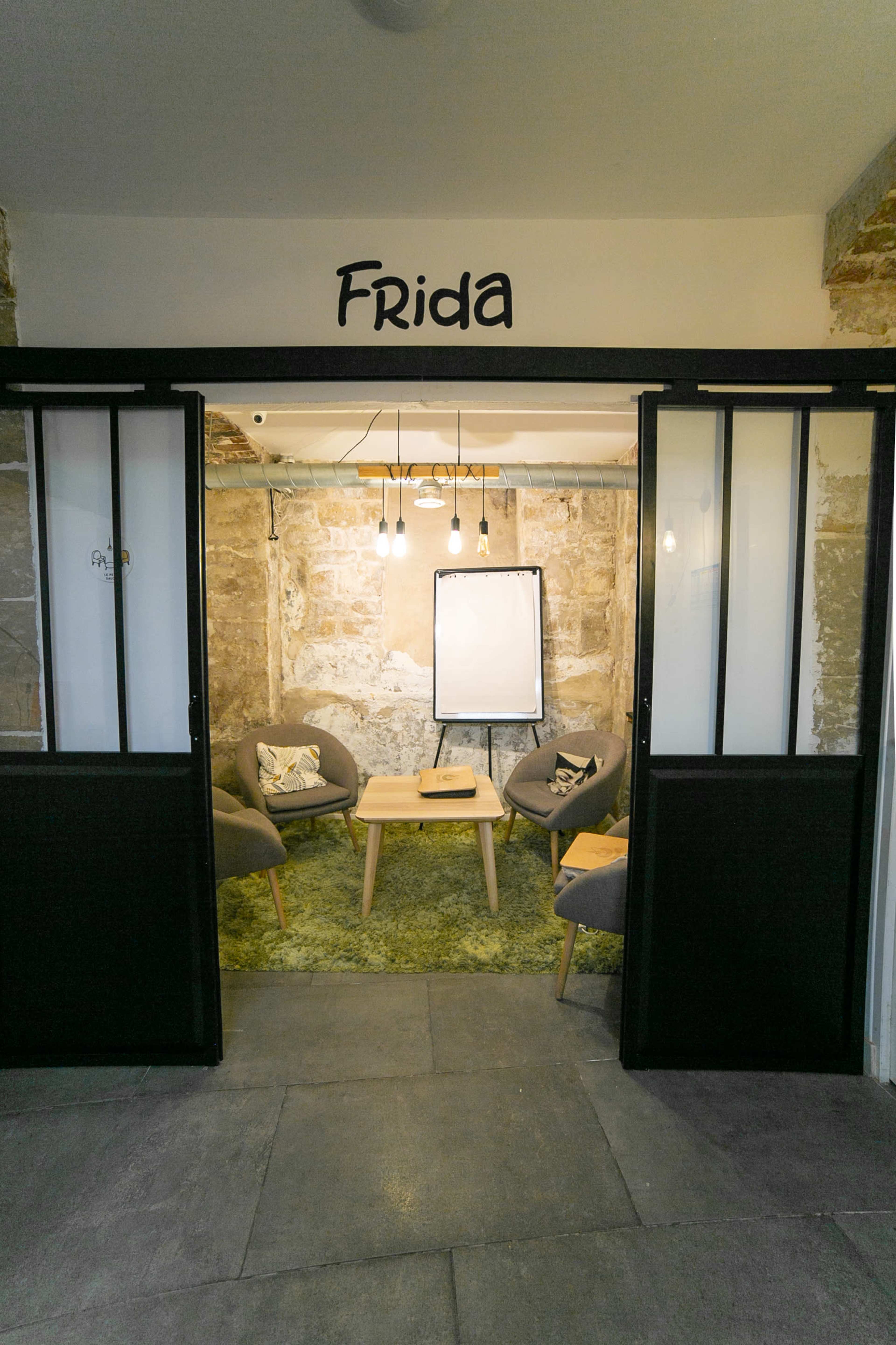 A cozy meeting space named "Frida" features four armchairs, a small table, and a whiteboard against a stone wall.